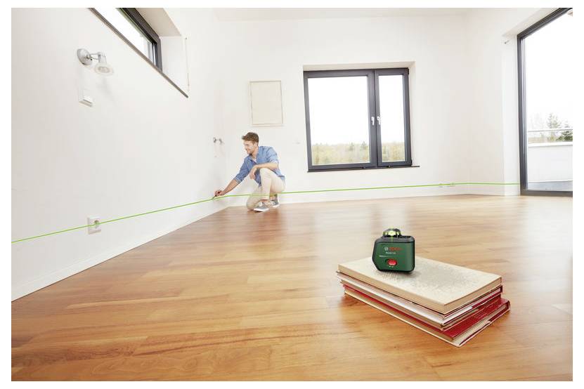 Arbeiter, der einen Raum mit einem Laser-Nivellier misst, grüne Linie an der Wand, während Bodenbelagsmaterialien im Vordergrund liegen.