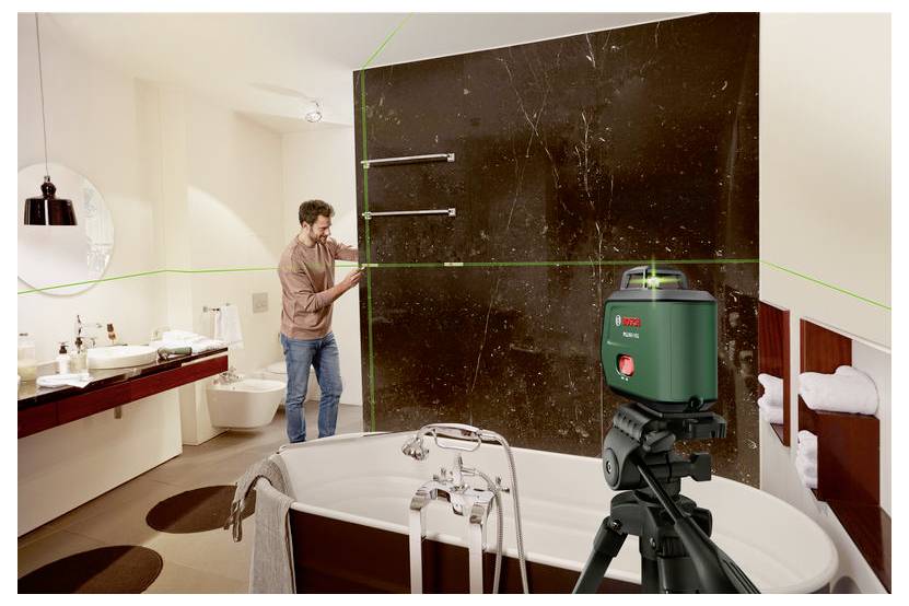 Ein Mann richtet ein großes dunkles Wandpanel in einem Badezimmer aus, wobei ein Laserniveau auf einem Stativ Hilfslinienprojektionen über eine Badewanne und ein Waschbecken wirft.