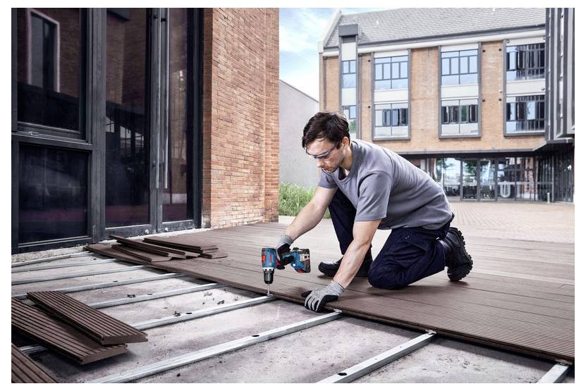 Ein Mann kniet auf einer Terrasse und befestigt mit einem Elektroschrauber Holzdeckelbretter über einem Metallrahmen neben einem modernen Gebäude.