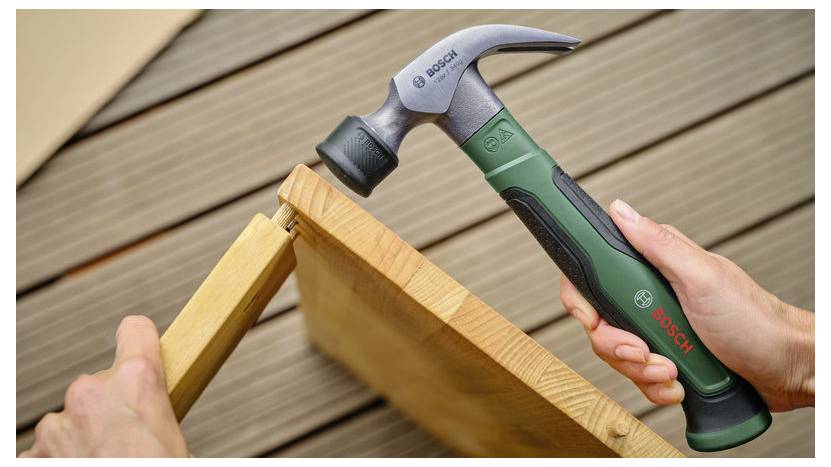 Hände verwenden einen Bosch-Krallenhammer, um eine Holzrahmen-Verbindung auf einer Terrasse herauszutreiben, wobei Reparaturarbeiten am Holzrahmen gezeigt werden.