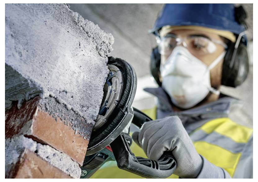 Arbeiter mit Schutzhelm, Schutzbrille, Maske und Handschuhen benutzt einen Elektroschleiffer, um eine Ziegelmauer zu schneiden oder zu glätten.