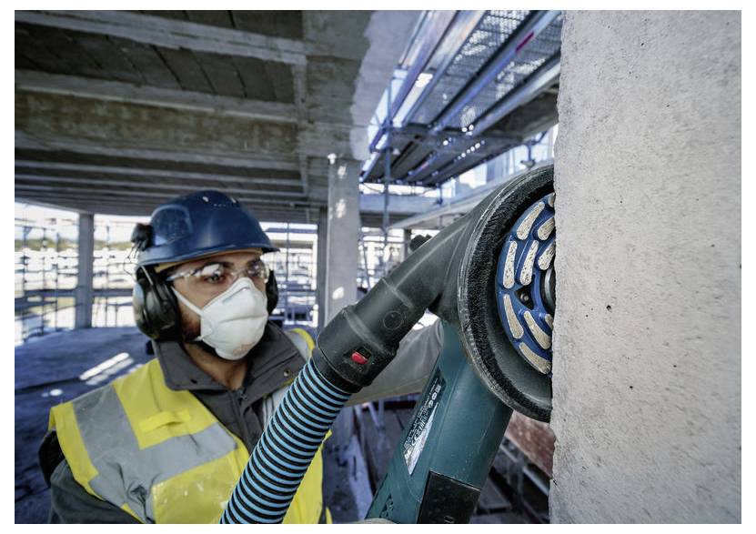 Bauarbeiter mit Schutzhelm, Sicherheitsweste und Maske, der ein Elektrowerkzeug auf einer Betonoberfläche auf einem Industriegelände verwendet.