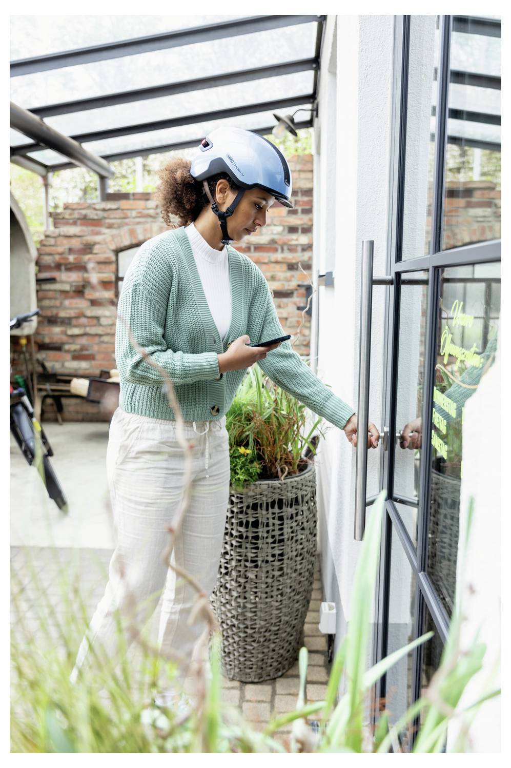 Eine Frau mit blauem Helm nutzt ein Smartphone, während sie eine Glastür auf einer Terrasse oder einem Balkon öffnet.