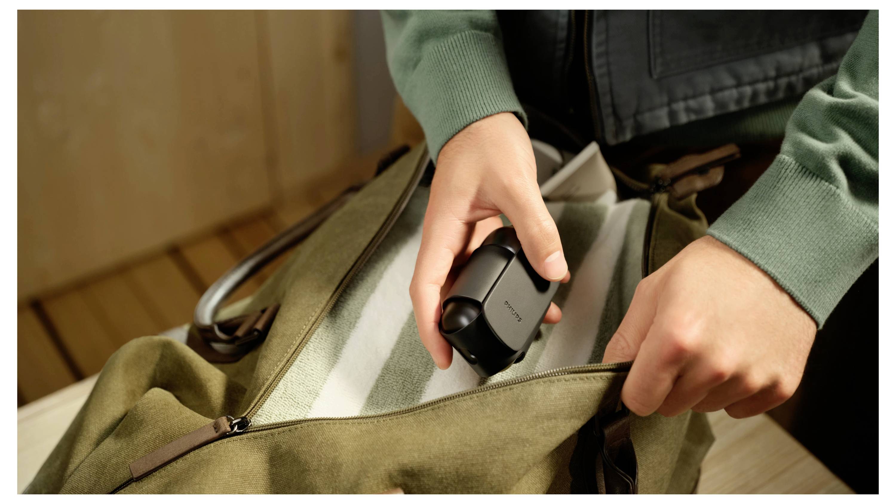 Eine Person packt ein kleines schwarzes Gerät in eine offene grüne Reisetasche auf einem Tisch.