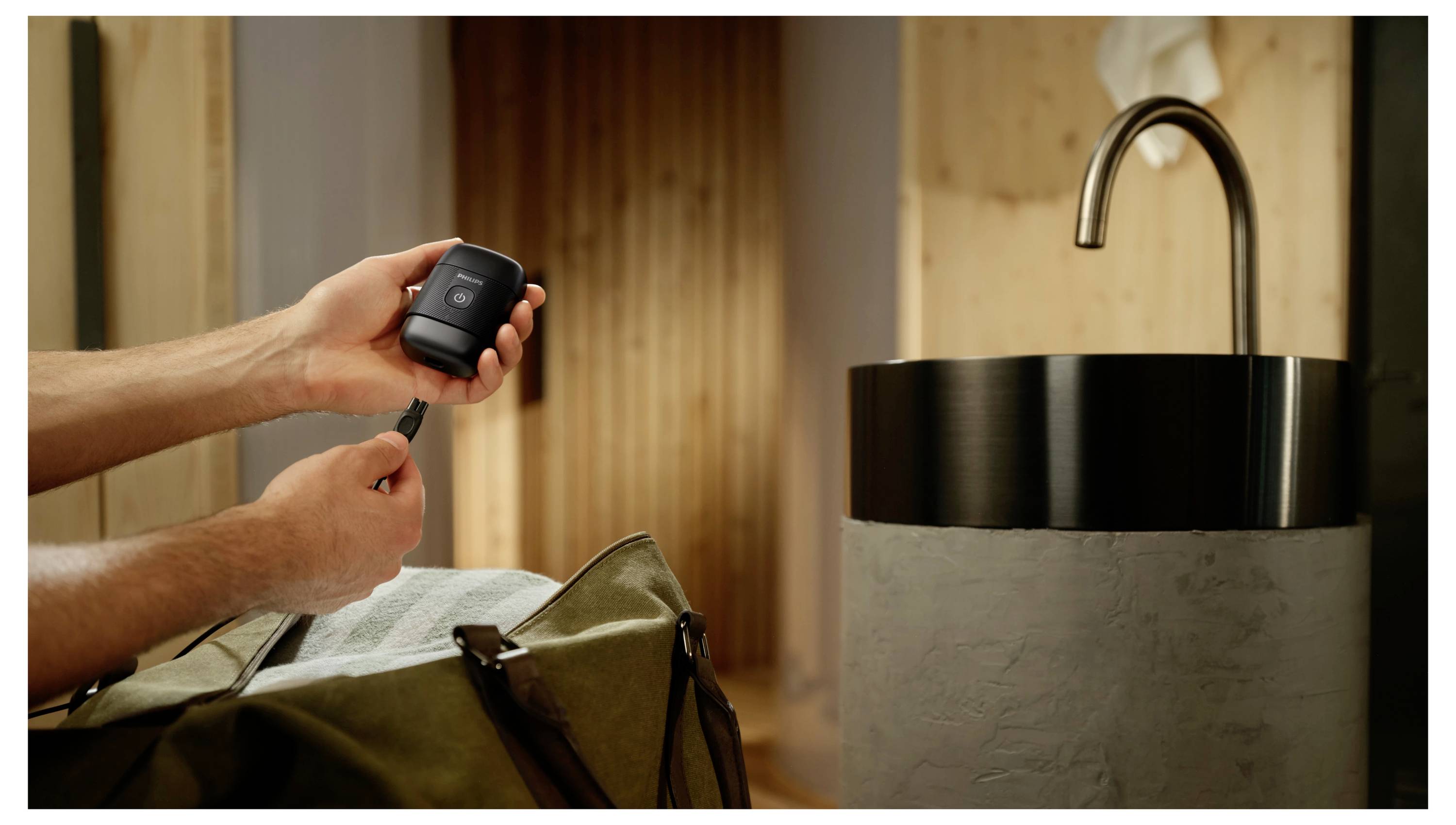 Eine Person lädt ein kleines Gerät mit einem Kabel neben einer offenen Tasche und einem Waschbecken in einem modernen Zimmer auf.