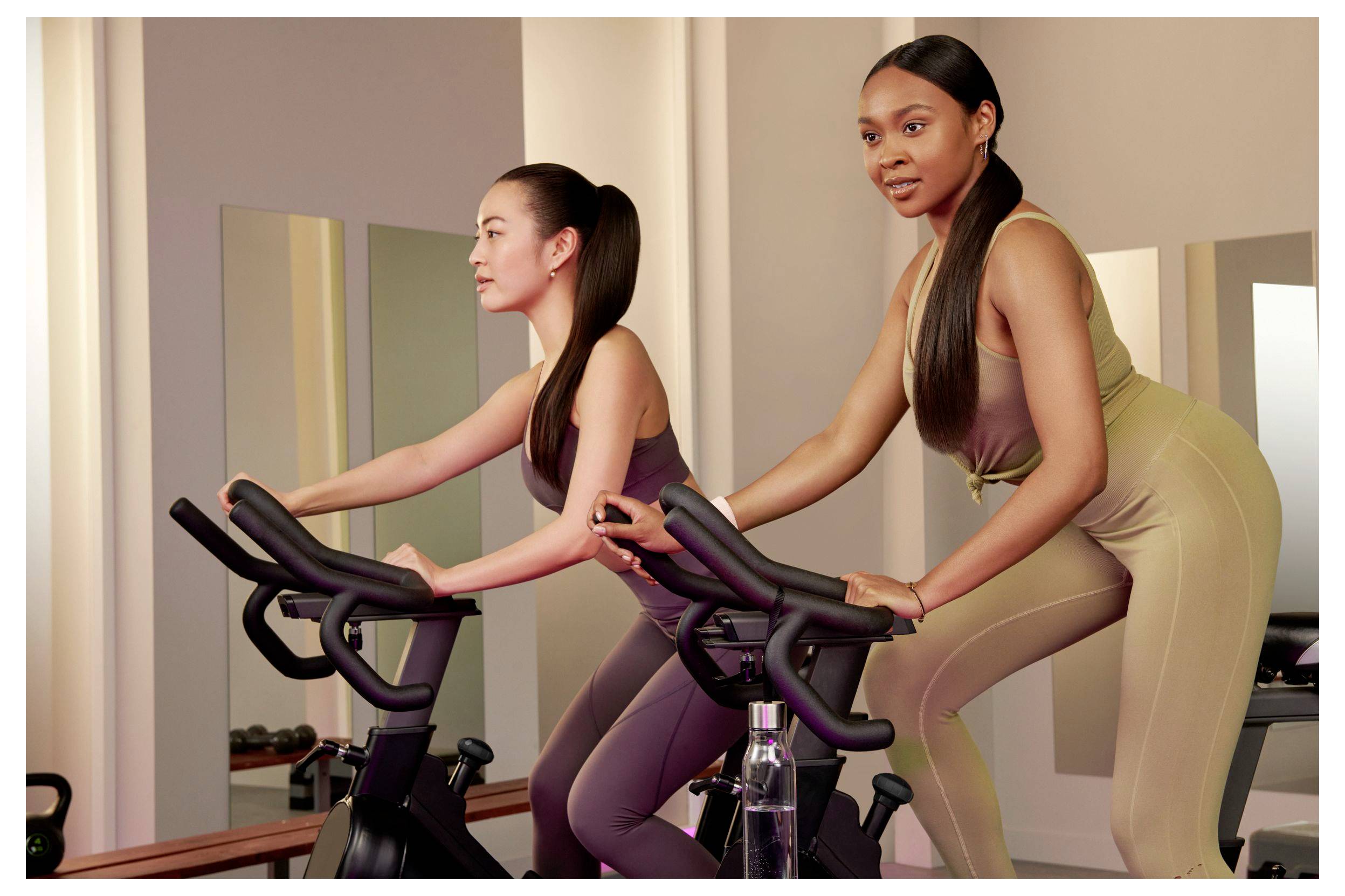 Zwei Frauen trainieren auf stationären Fahrrädern in einem Gym-Studio, eine vorne und eine hinten, beide lehnen sich nach vorne und treten in die Pedale.