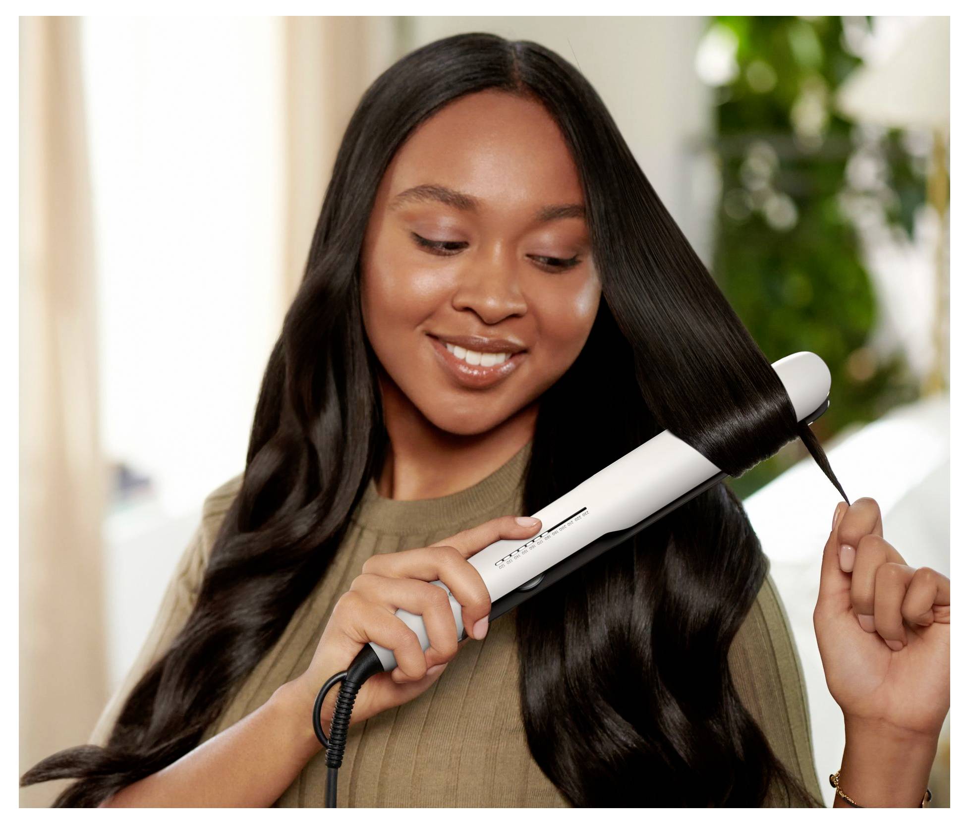 Lächelnde Frau richtet langes dunkles Haar mit einem weißen Styling-Gerät auf und hält eine Haarsträhne in einer hellen Innenumgebung.