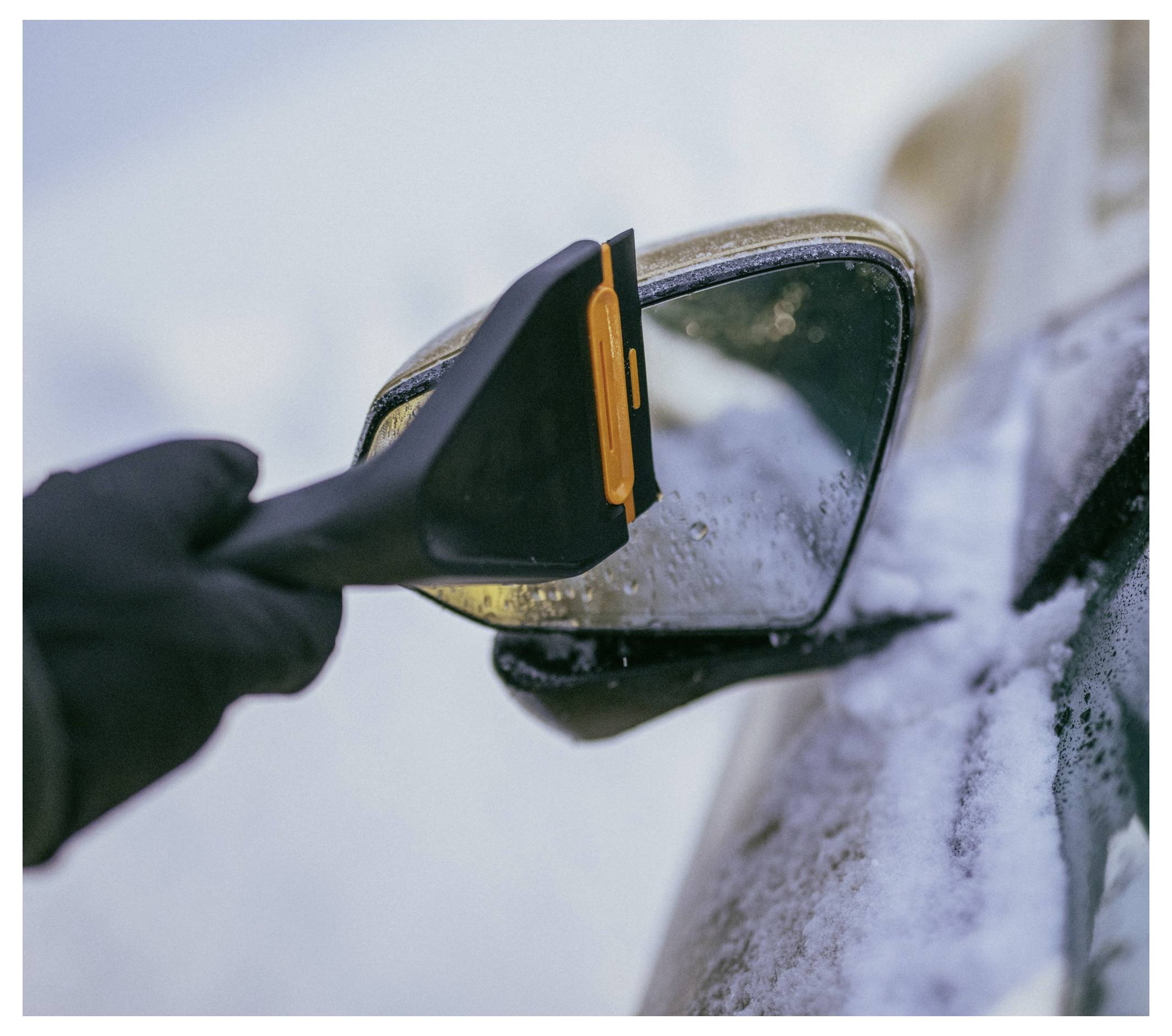Mit behandschuhter Hand wird Eis mit einem Eiskratzer von einem Außenspiegel eines schneebedeckten Fahrzeugs abgekratzt.