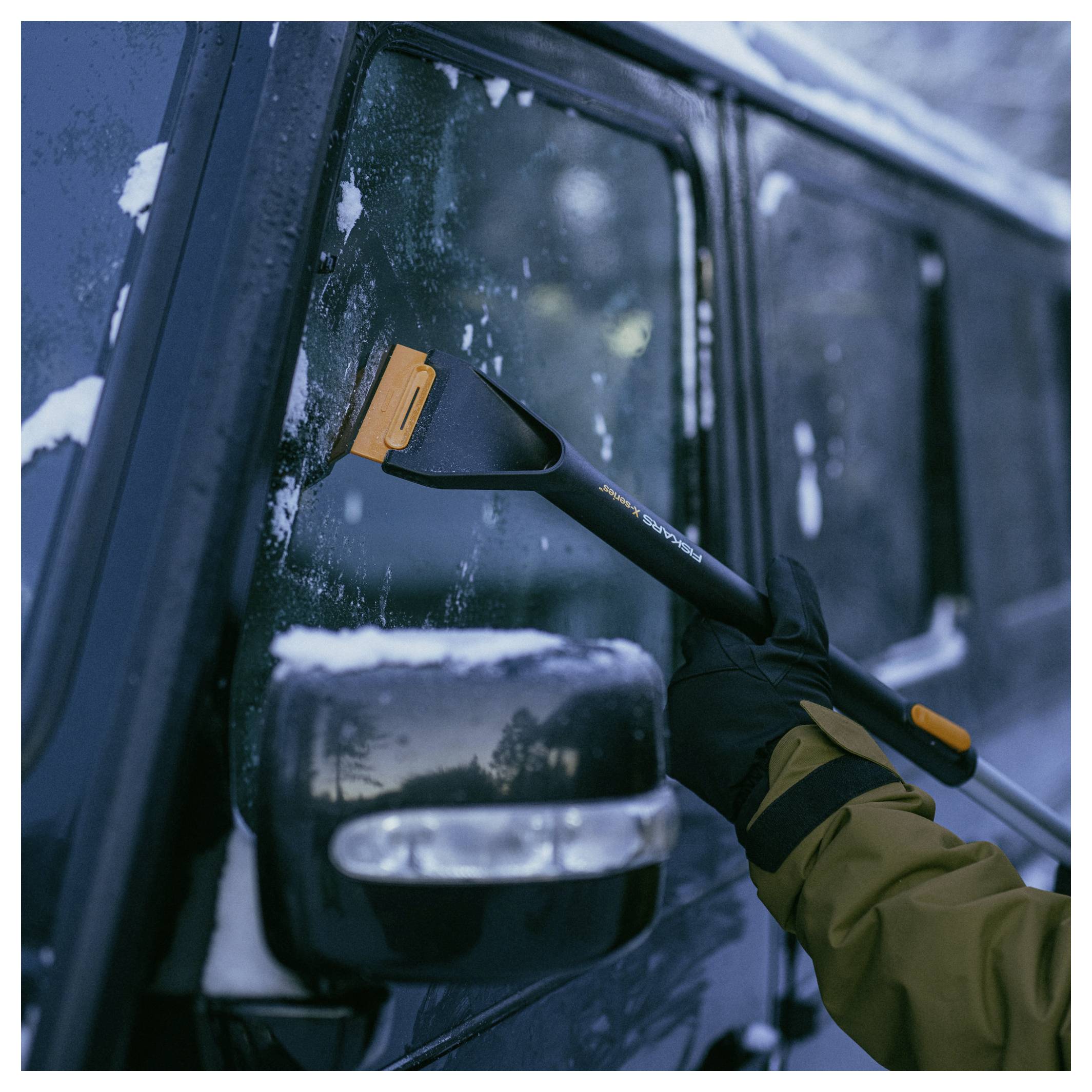 Handschuh tragende Hand, die eine Schneebürste verwendet, um Schnee und Eis von einem Fahrzeugfenster und Seitenspiegel zu kratzen.