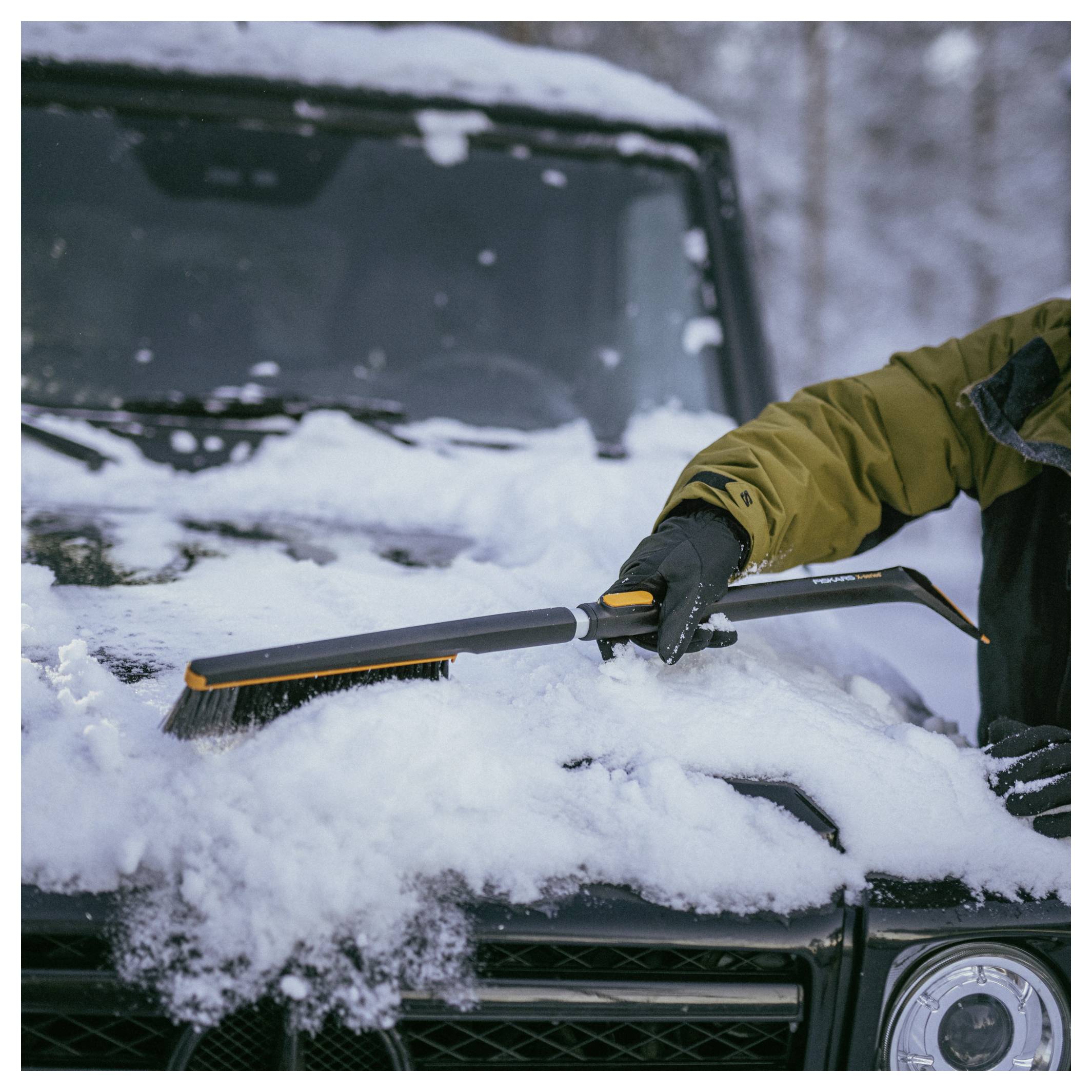 Person in Winterjacke, die mit einer Bürste Schnee von der Windschutzscheibe und der Motorhaube eines geparkten Autos räumt.
