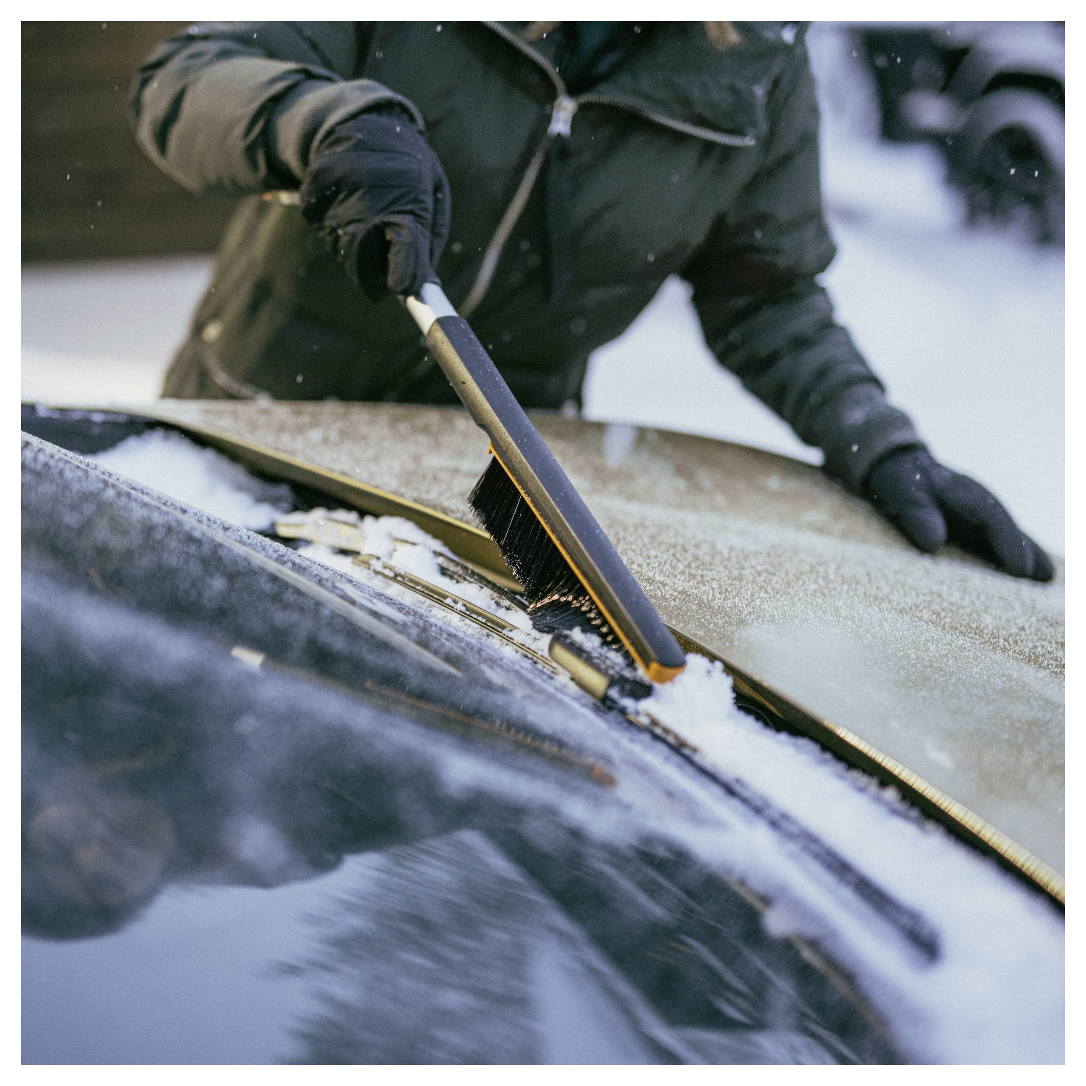 Eine Person, die Schnee und Eis mit einer tragbaren Bürste von einer Autoscheibe im Freien im Winter entfernt.