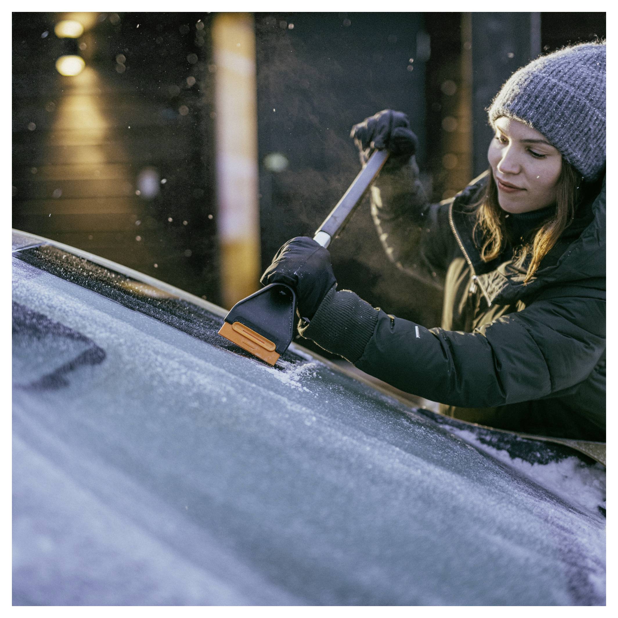 Frau in Winterkleidung, die mit einem Eiskratzer Reif von einer Autoscheibe kratzt