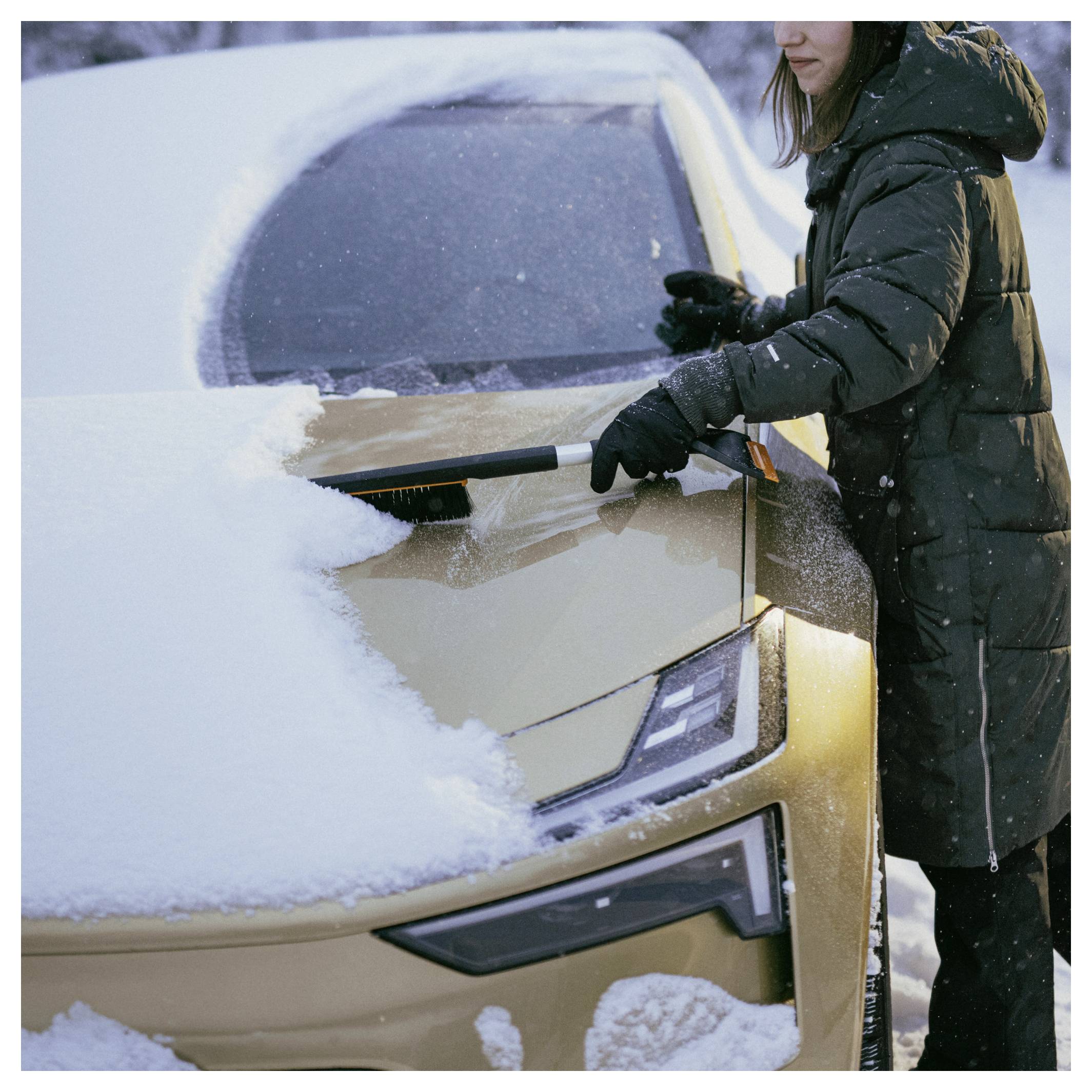 Person in einem Wintermantel, die Schnee von einer Autoscheibe und Motorhaube in einer verschneiten Szene abbürrstet.