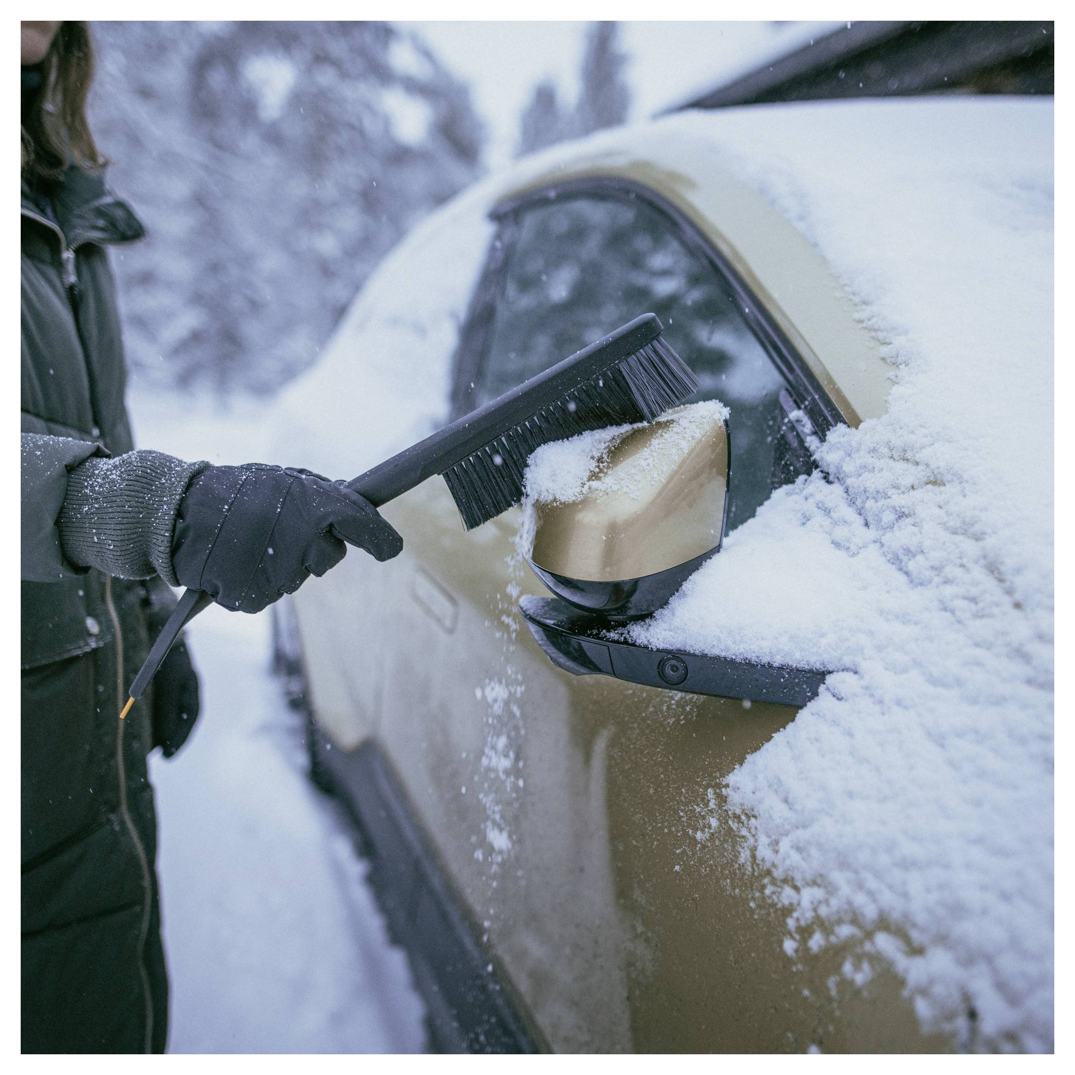 Behandschuhte Hand, die eine Schneebürste/einen Eiskratzer verwendet, um Frost und Schnee von einer Autoscheibe zu entfernen.