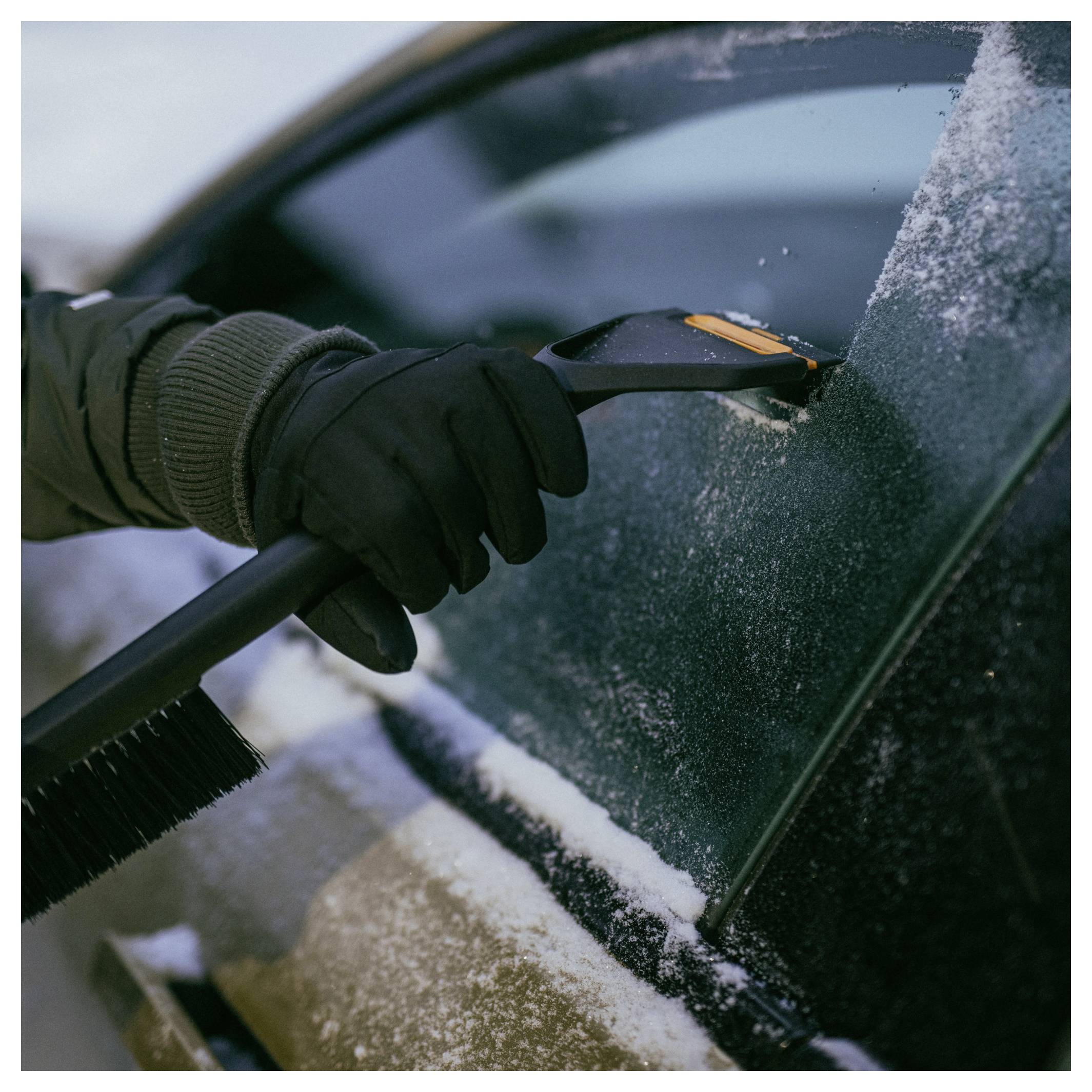 Behandschuhte Hand, die einen Eiskratzer verwendet, um Reif und Schnee von einer Autoscheibe zu entfernen.