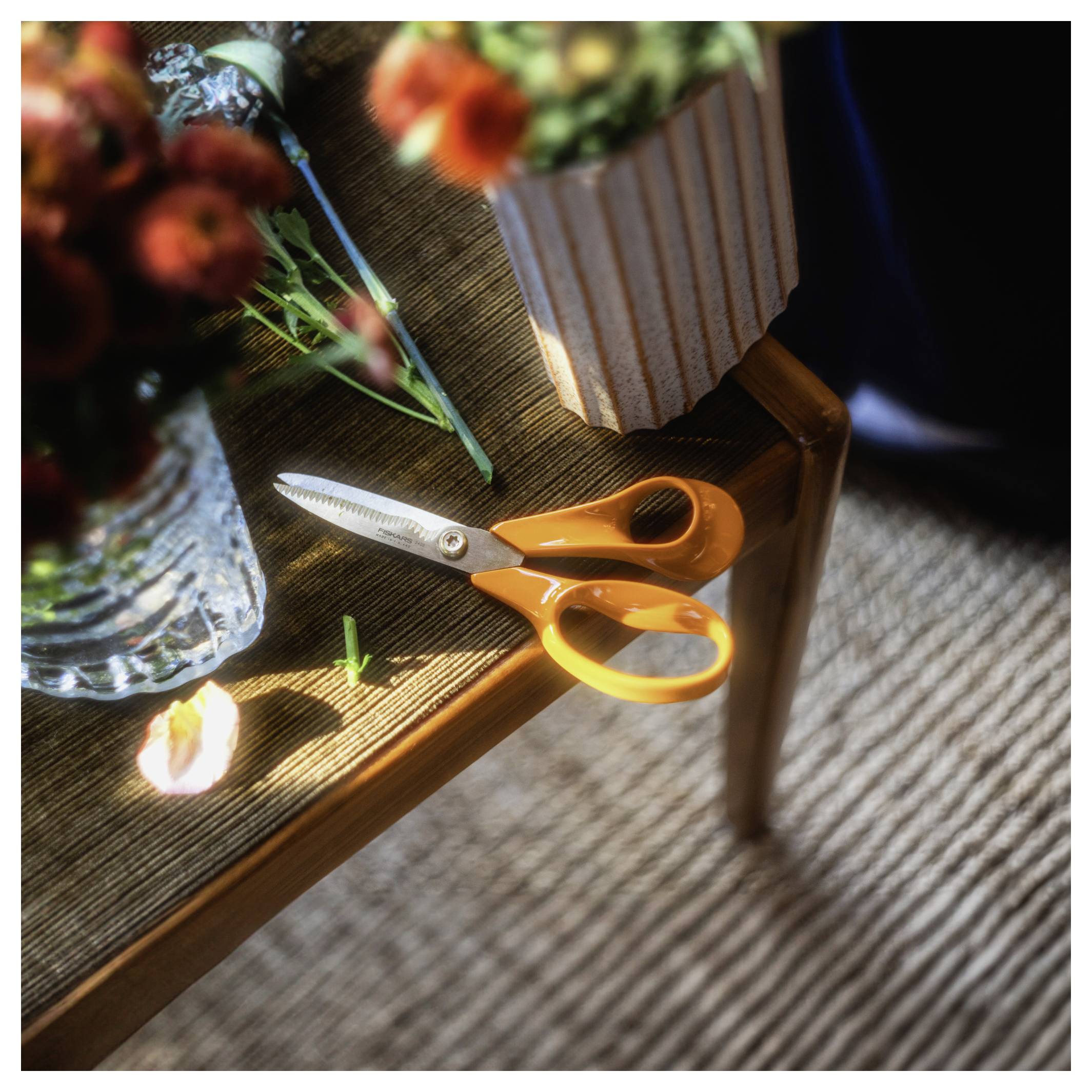 Orange-farbige Schere am Rand eines Holztisches neben einer Blumenvase, mit Sonnenlicht und einem gestreiften Teppich im Hintergrund.
