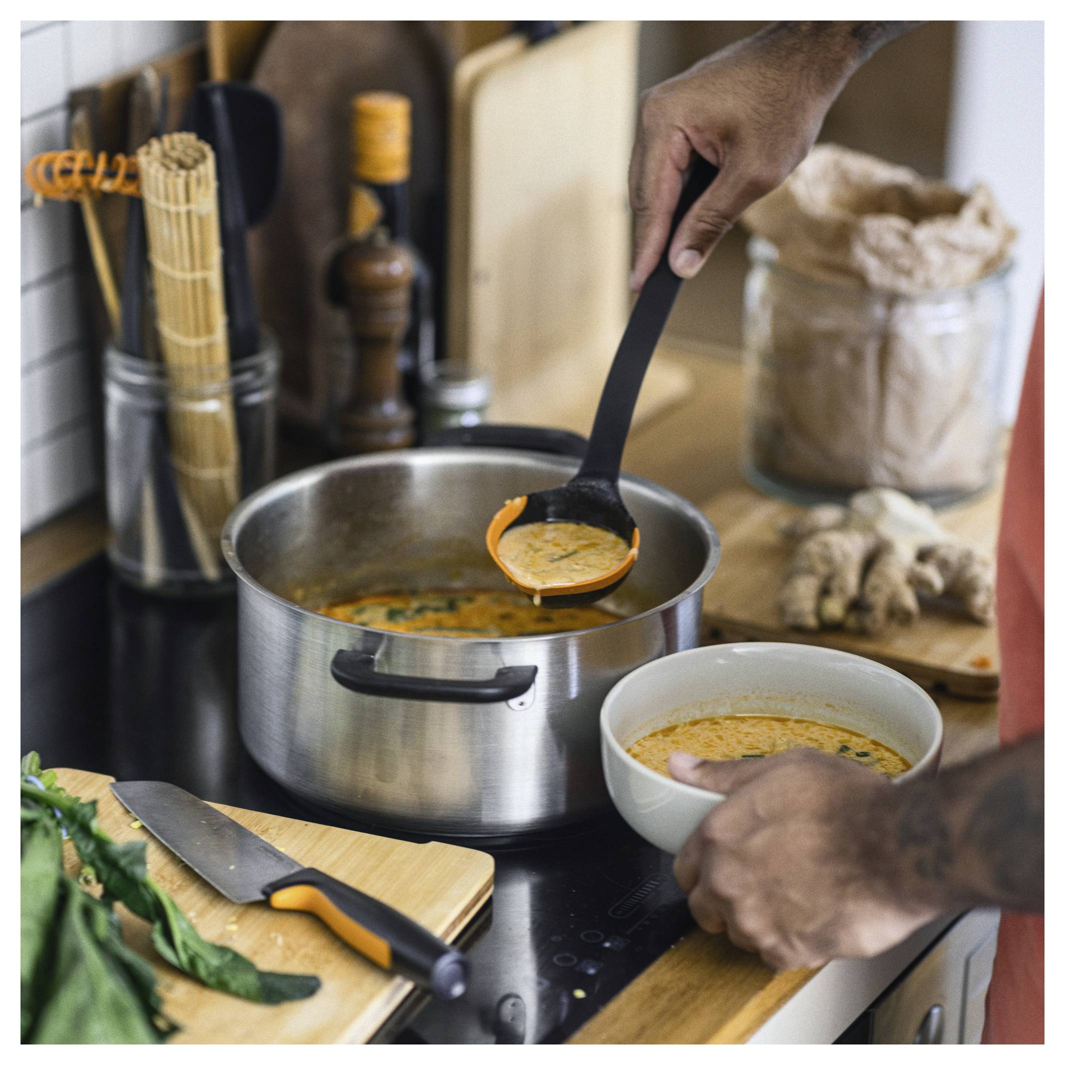 Eine Person schöpft Suppe mit einer Kelle aus einem Topf in eine Schüssel auf einem Küchenherd, mit Utensilien und Zutaten in der Nähe.