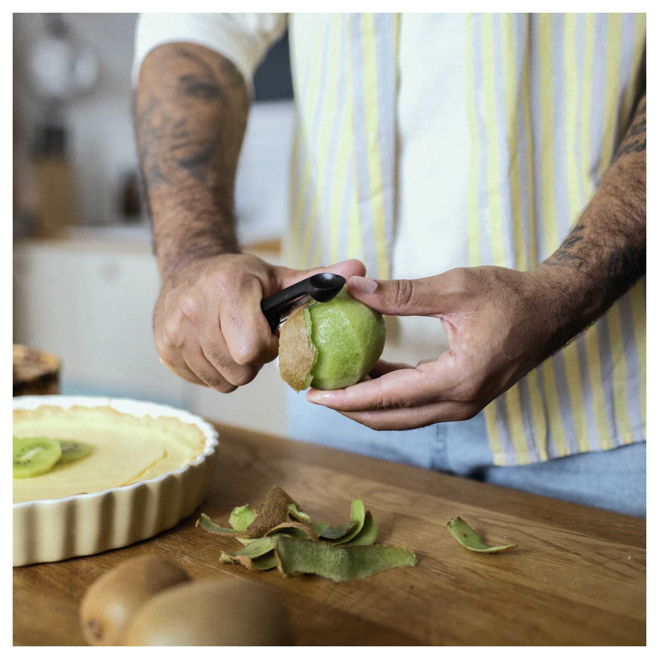 Person schält eine grüne Kiwi über eine Holzarbeitsplatte neben einer Obsttorte, mit Kiwischale und anderen Früchten in der Nähe.
