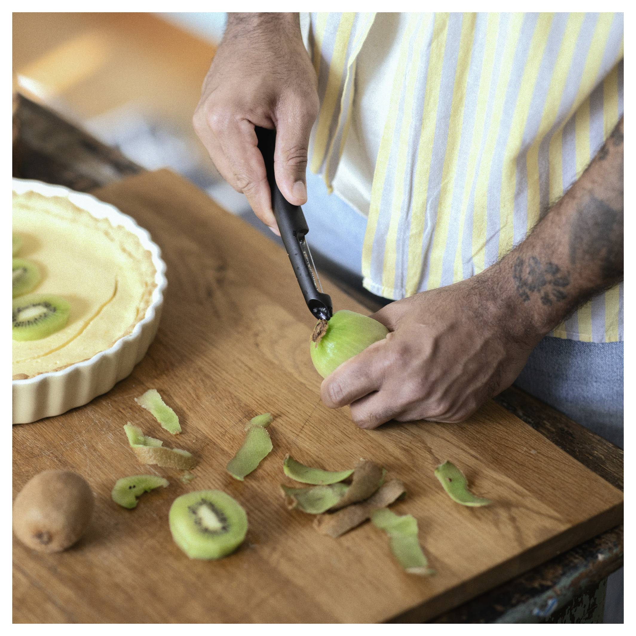Eine Person schält eine Kiwi auf einem Schneidebrett neben einer Kiwi-Torte oder einem Kiwi-Kuchen, mit verstreuten Schalen in der Nähe.