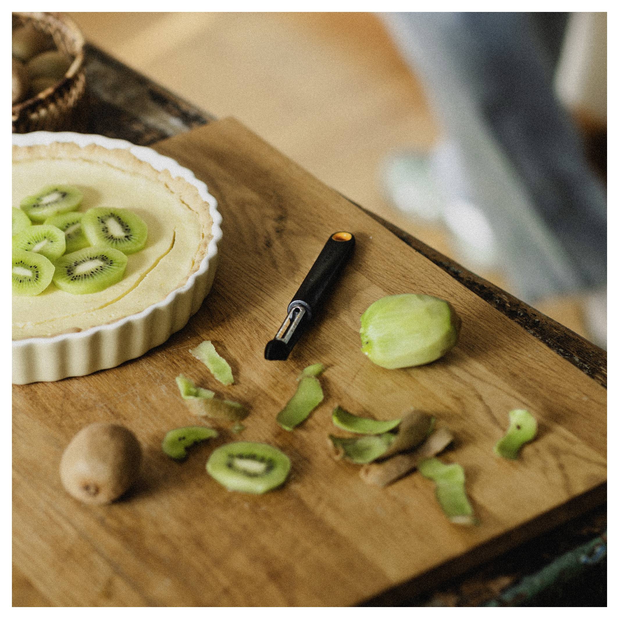 Kiwi-Tarte auf einem Holzschneidebrett mit geschälten und geschnittenen Kiwis, Kiwischalen und einem Schälmesser in der Nähe.