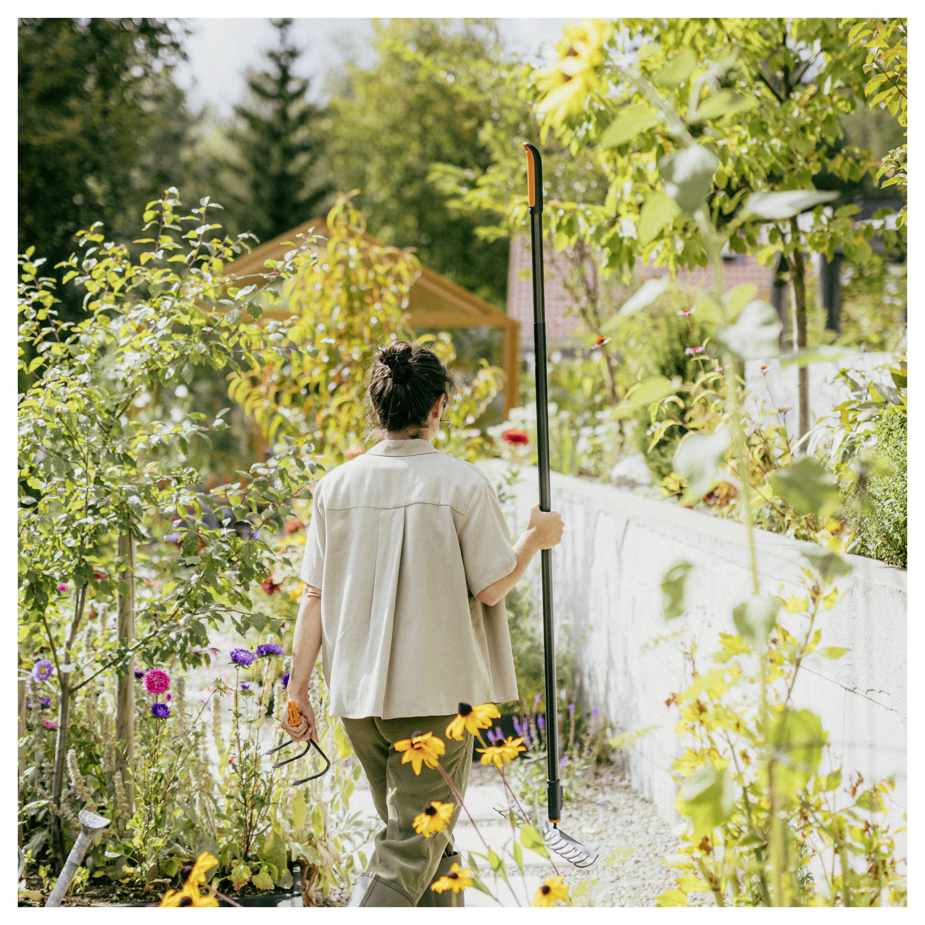 Person, die in einem üppigen Hintergarten arbeitet, hält ein Werkzeug mit langem Stiel neben einem blumenreichen Weg und einer weißen Wand in der Nähe eines kleinen Schuppens.