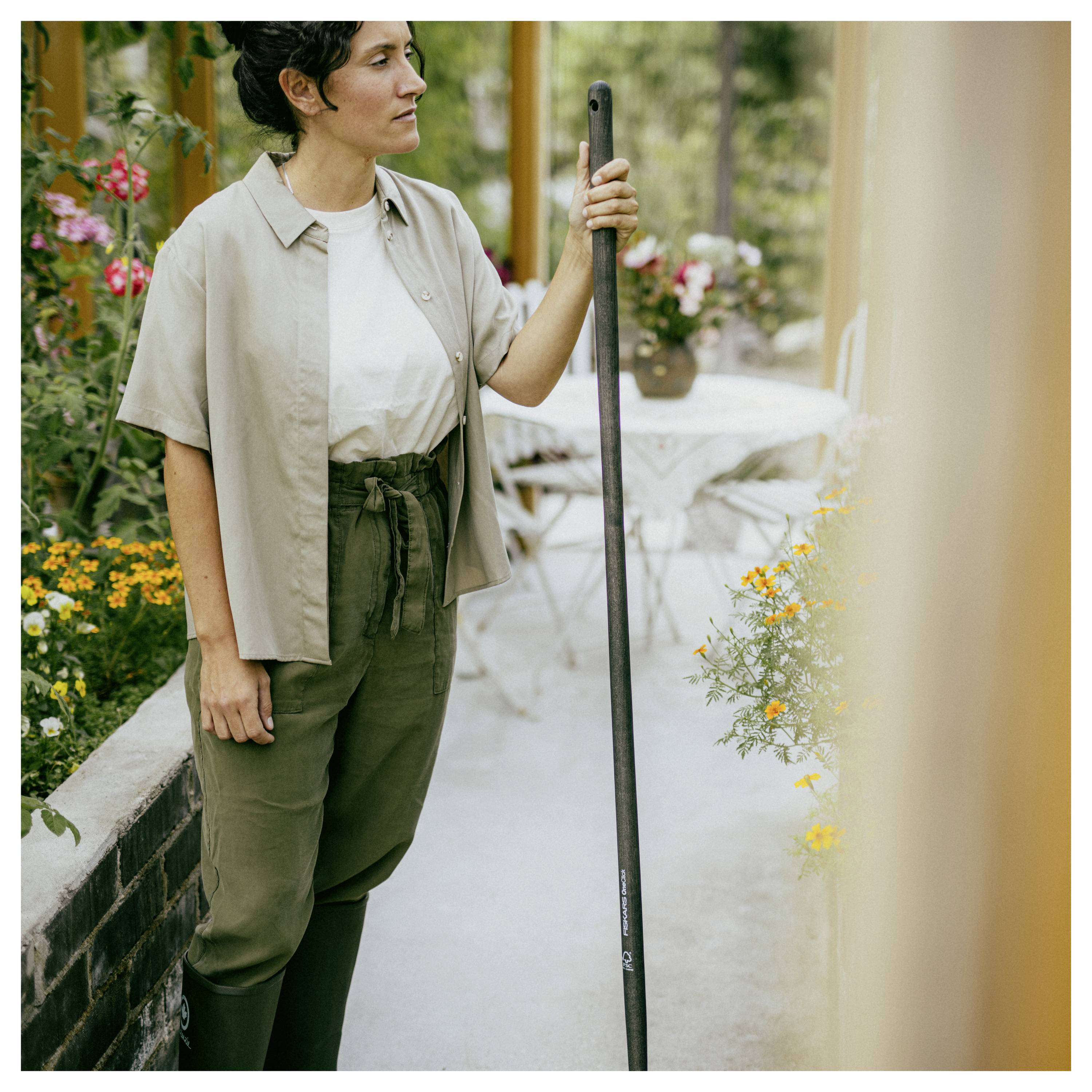 Person in Gartenkleidung steht in einem blumenreichen Innenhof und hält neben einem Terrassentisch und Stühlen eine lange Stange oder ein Werkzeug.