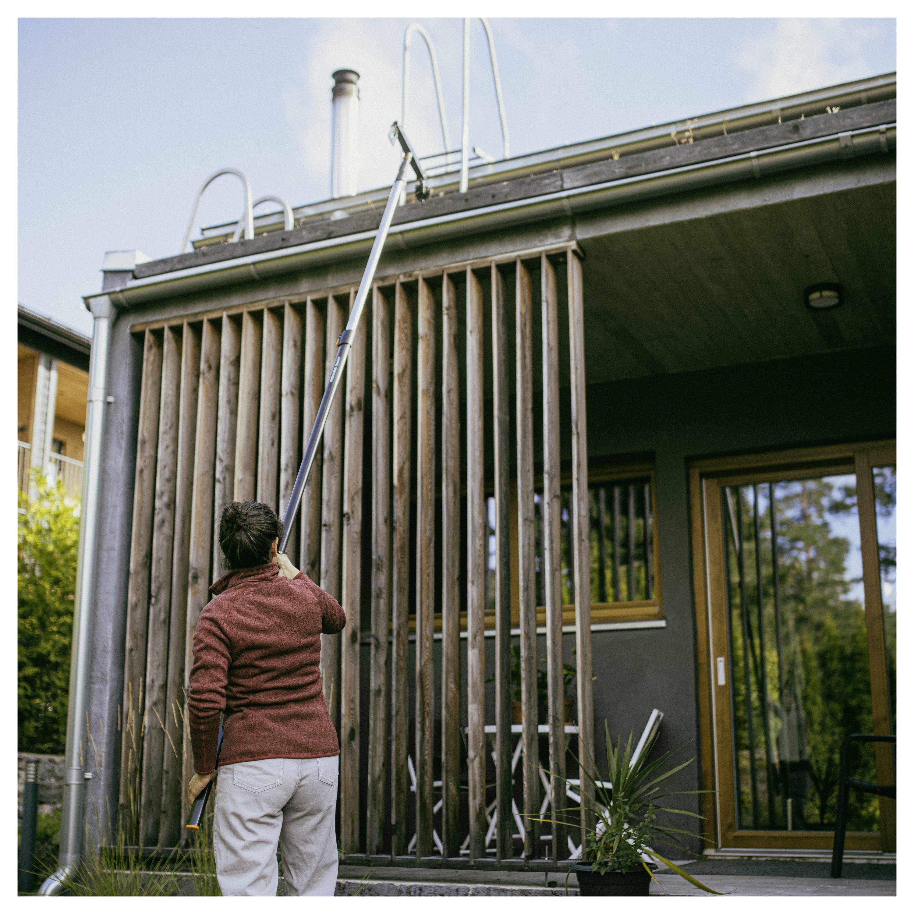 Person mit einer verlängerten Stange zur Reinigung des oberen Außenbereichs eines modernen Hauses mit vertikalen Holzlamellen und einem Terrassenbereich.