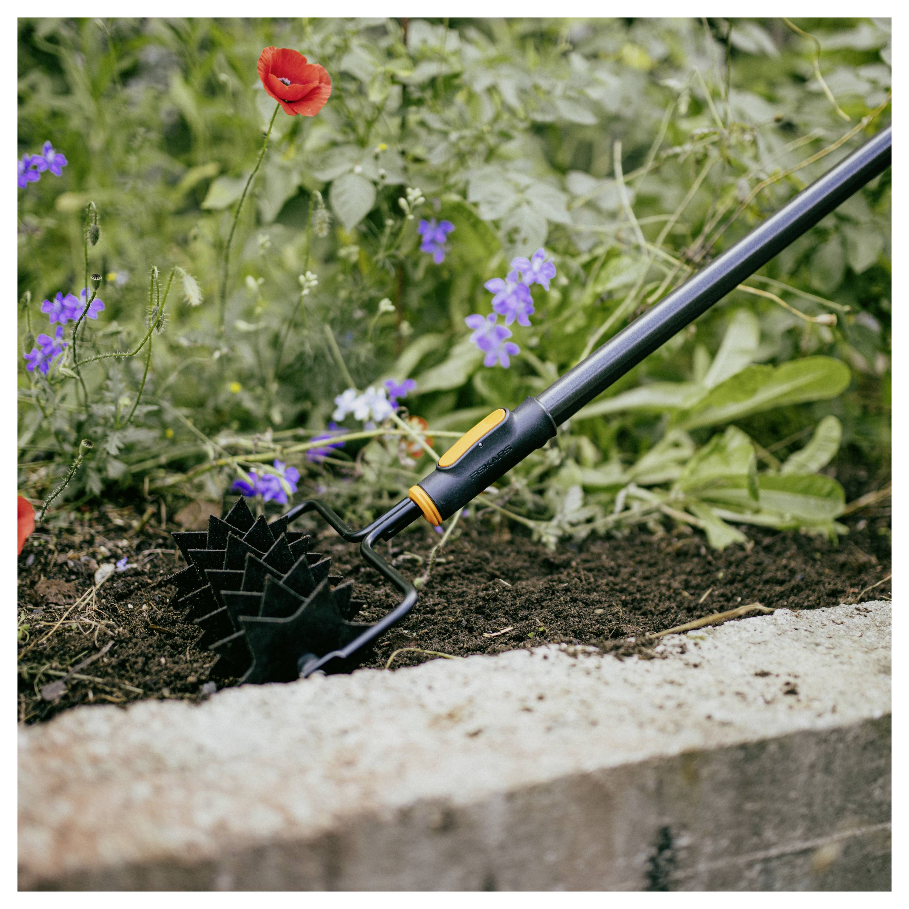 Gartenhandhacke mit schwarzem Griff ruht in der Erde neben blühenden Blumen und einer Betonbegrenzung.