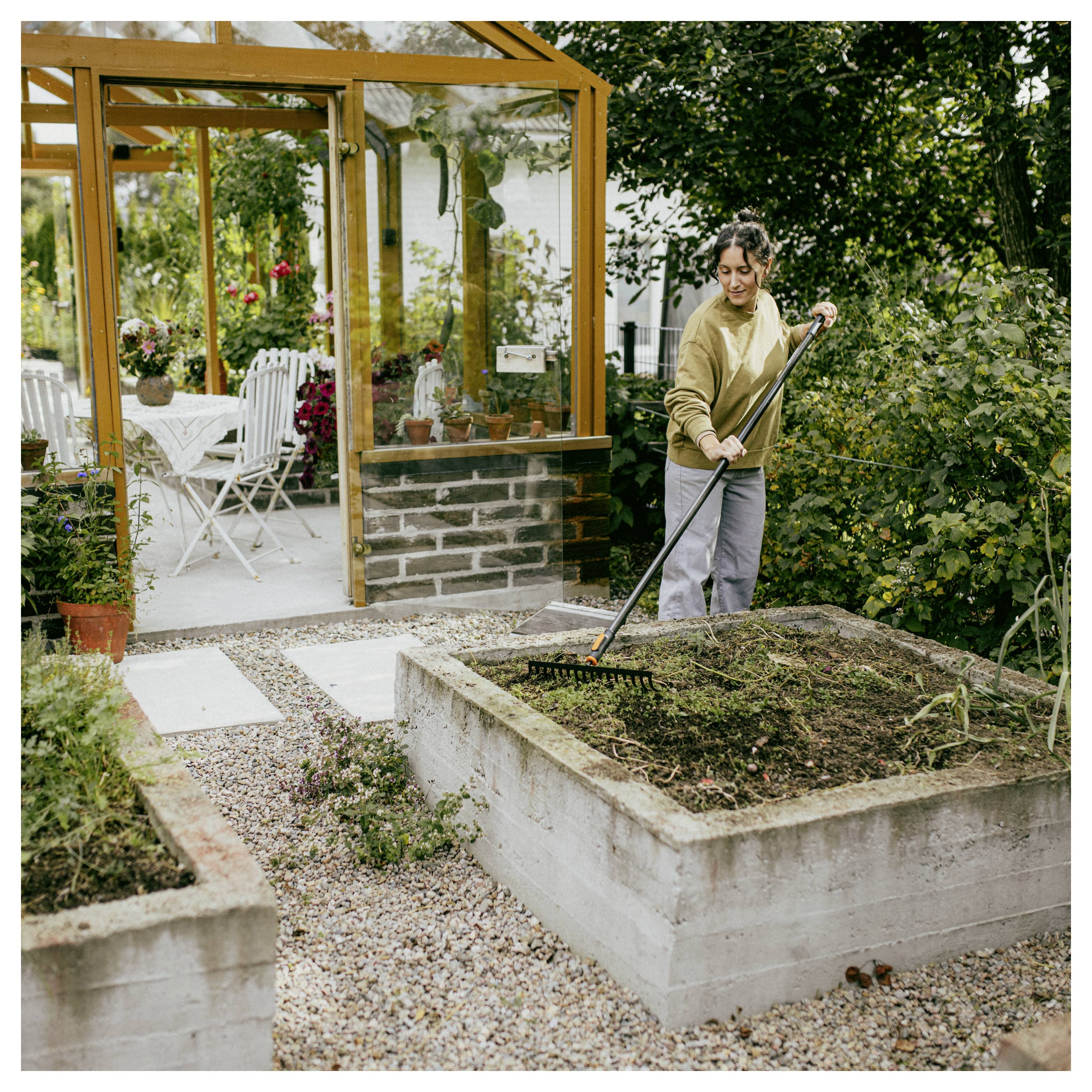 Eine Frau harkt ein erhöhtes Gartenbeet neben einem Gewächshaus und pflegt die Erde in einem Hinterhofgarten.