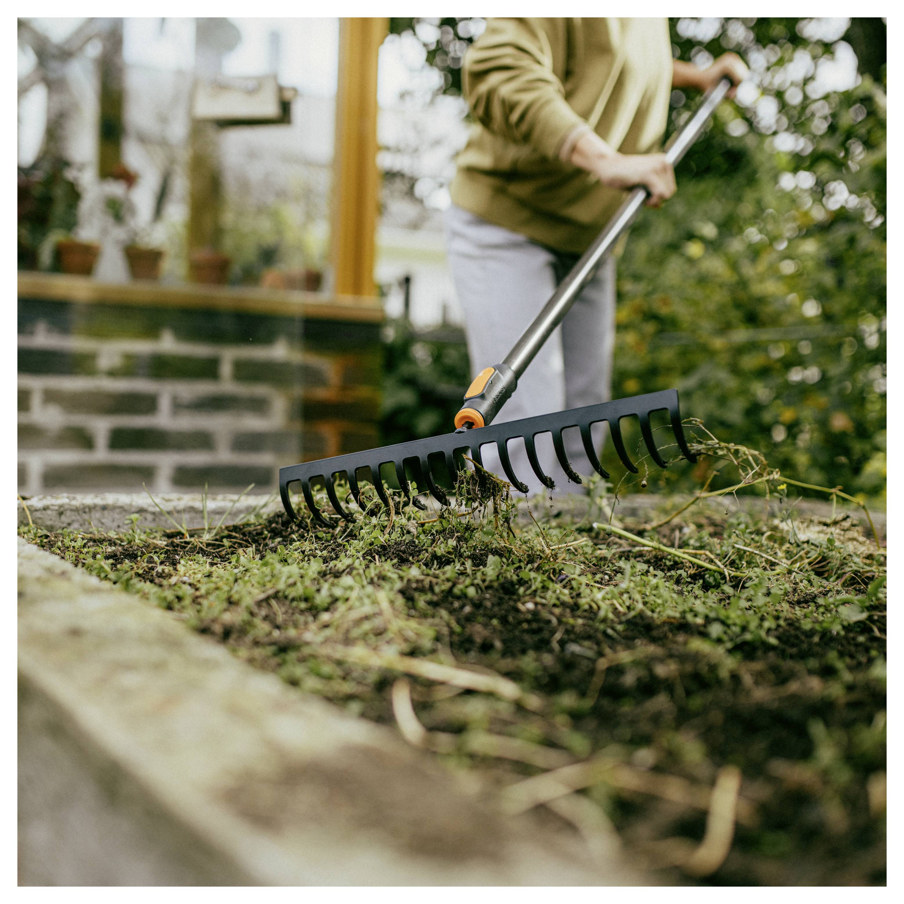 Eine Person nutzt einen Gartenrechen, um Blätter und Schmutz von einem Hof neben einem Haus zu entfernen.