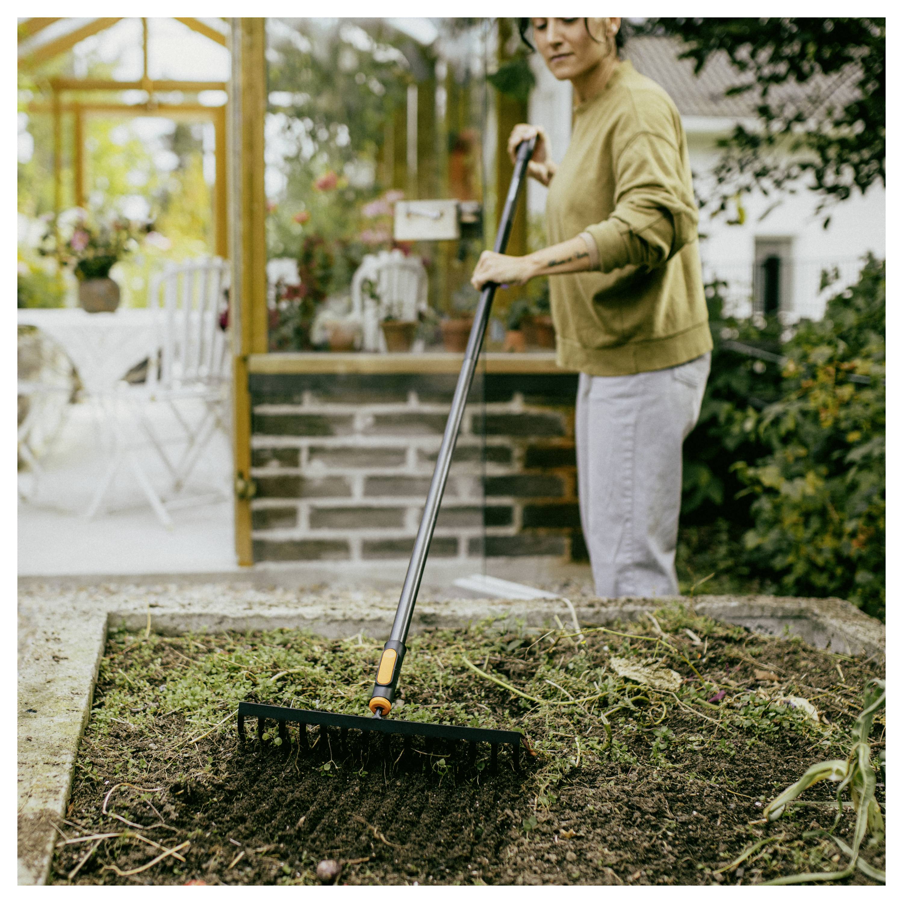Person, die ein kleines Gartenbeet im Hinterhof harkt, Erde und Unkraut neben einem Gewächshaus entfernt.