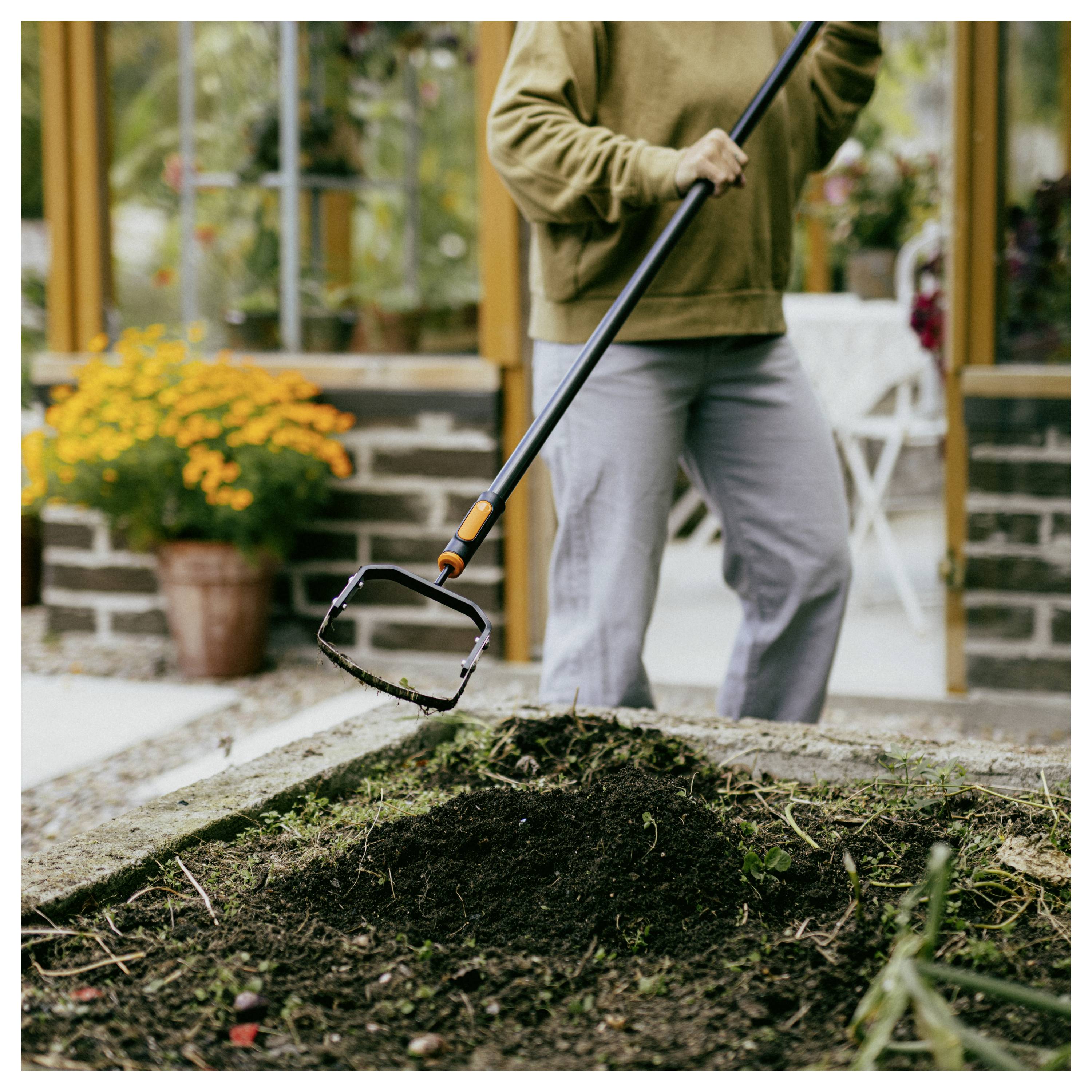 Eine Person mit einem langstieligen Gartenwerkzeug, die Unkraut oder Schutt aus einem erhöhten Outdoor-Blumenbeet vor einem Haus entfernt.