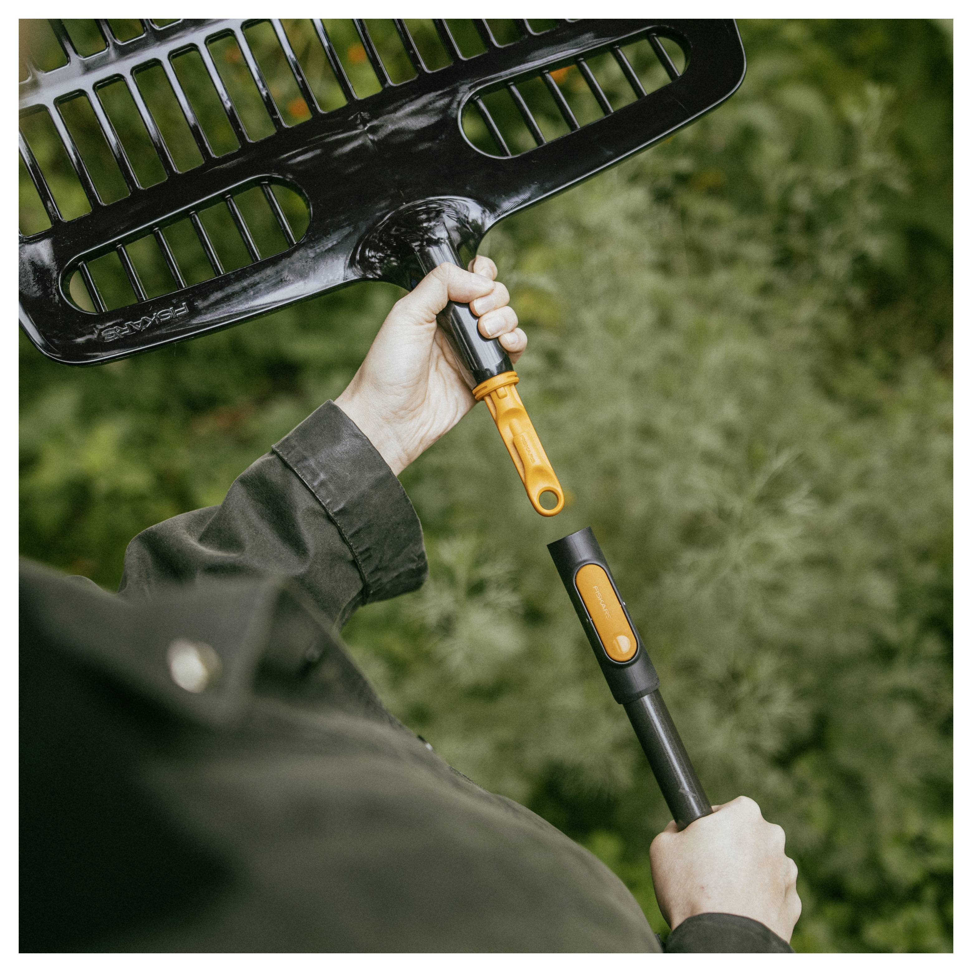 Eine Person hält eine große schwarze Gartenharke mit orangefarbenem Griff vor einem unscharfen grünen Außenhintergrund.