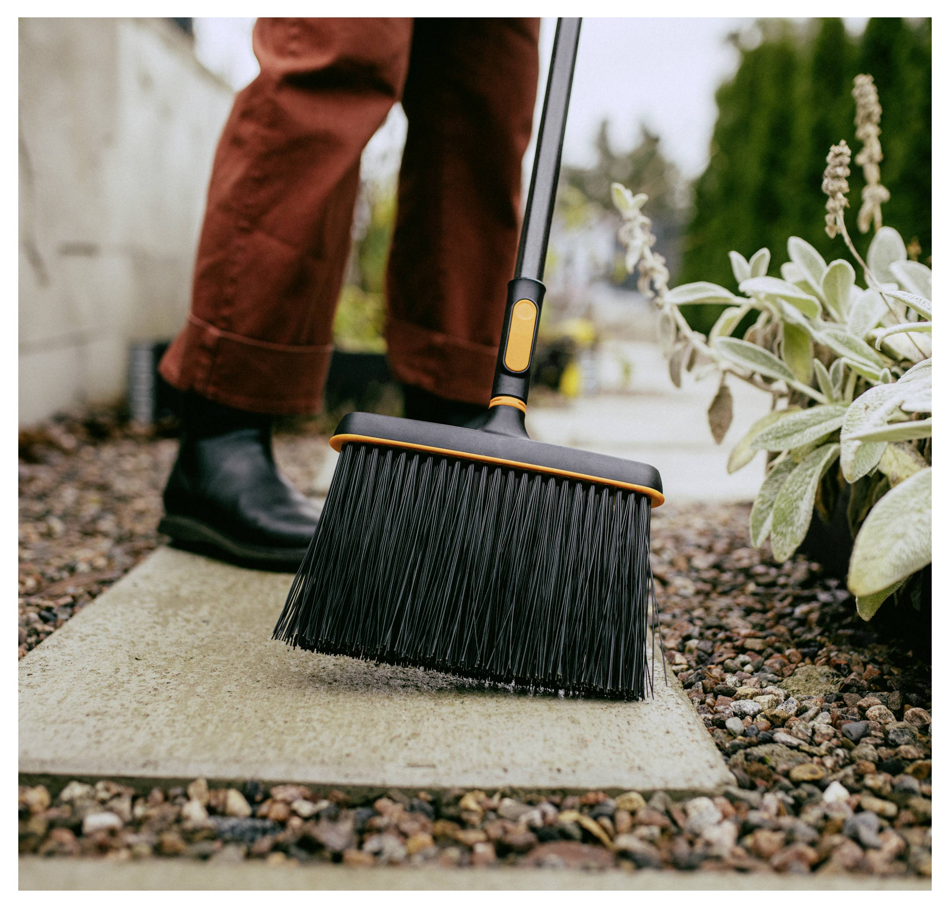 Nahaufnahme einer Person, die eine Steinterrasse mit einem schwarzen Besen neben Gartenpflanzen fegt, wobei Außenkies und Pflasterung gereinigt werden.