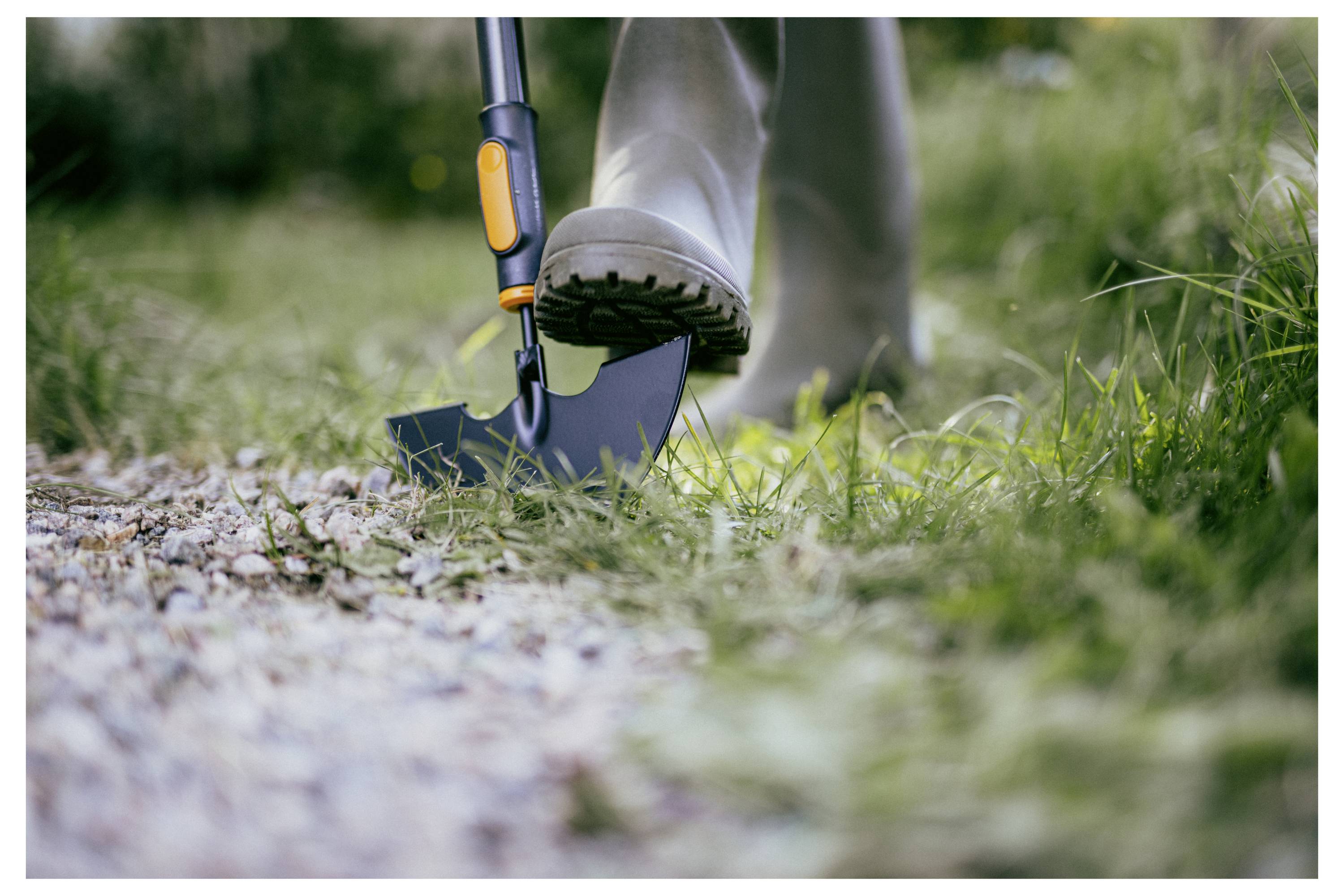Nahaufnahme einer Person, die mit einem Spaten entlang eines Kieswegs in grasbewachsenem Boden gräbt, wobei ein Stiefel in der Nähe der Klinge drückt.