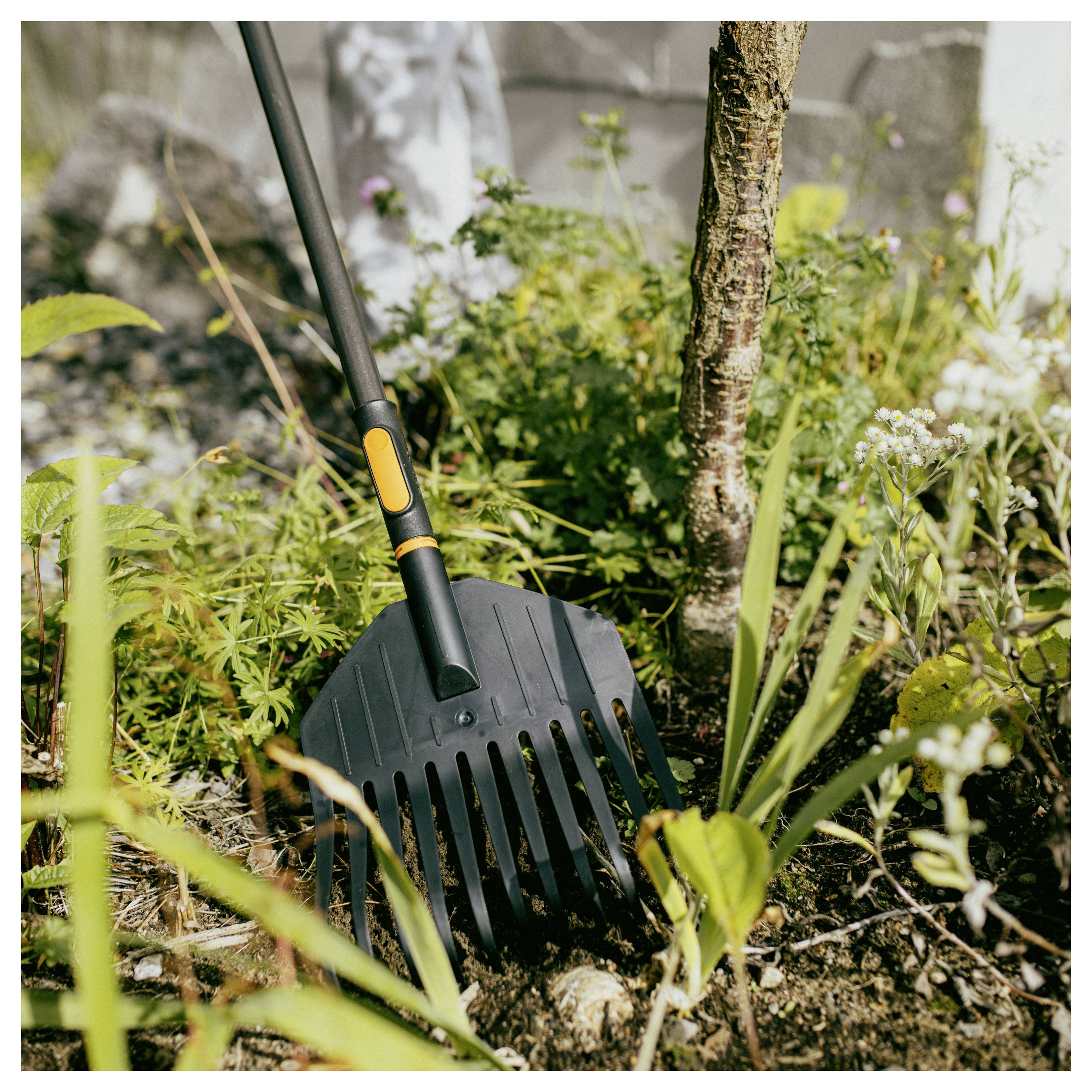 Gartenrechen lehnt in einem bepflanzten Blumenbeet mit Erde, Gräsern und einem kleinen Baumstamm im Hintergrund.