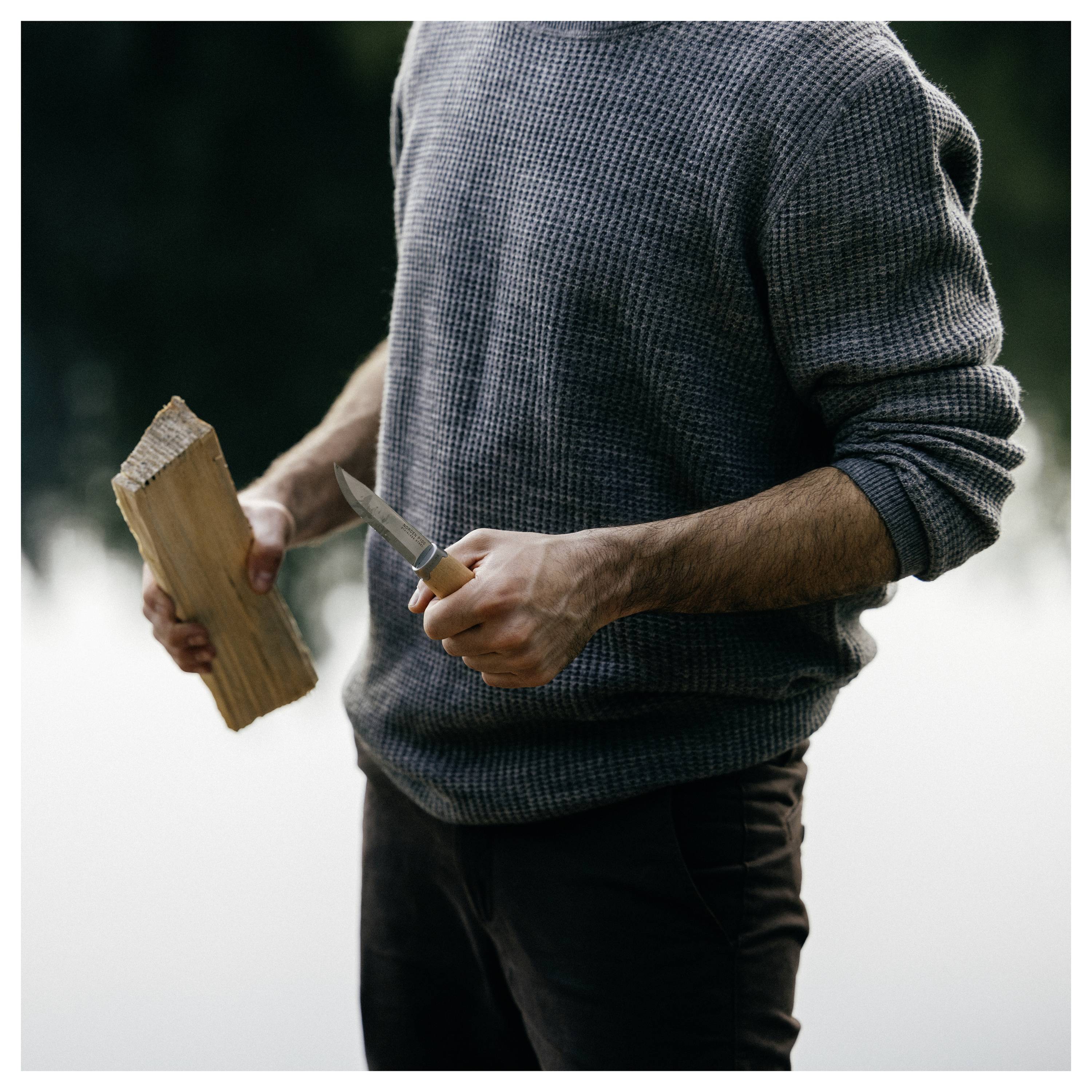 Person in einem grauen Pullover, der ein Stück Holz und ein Messer hält und sich darauf vorbereitet, im Freien zu schnitzen oder zu schneiden.