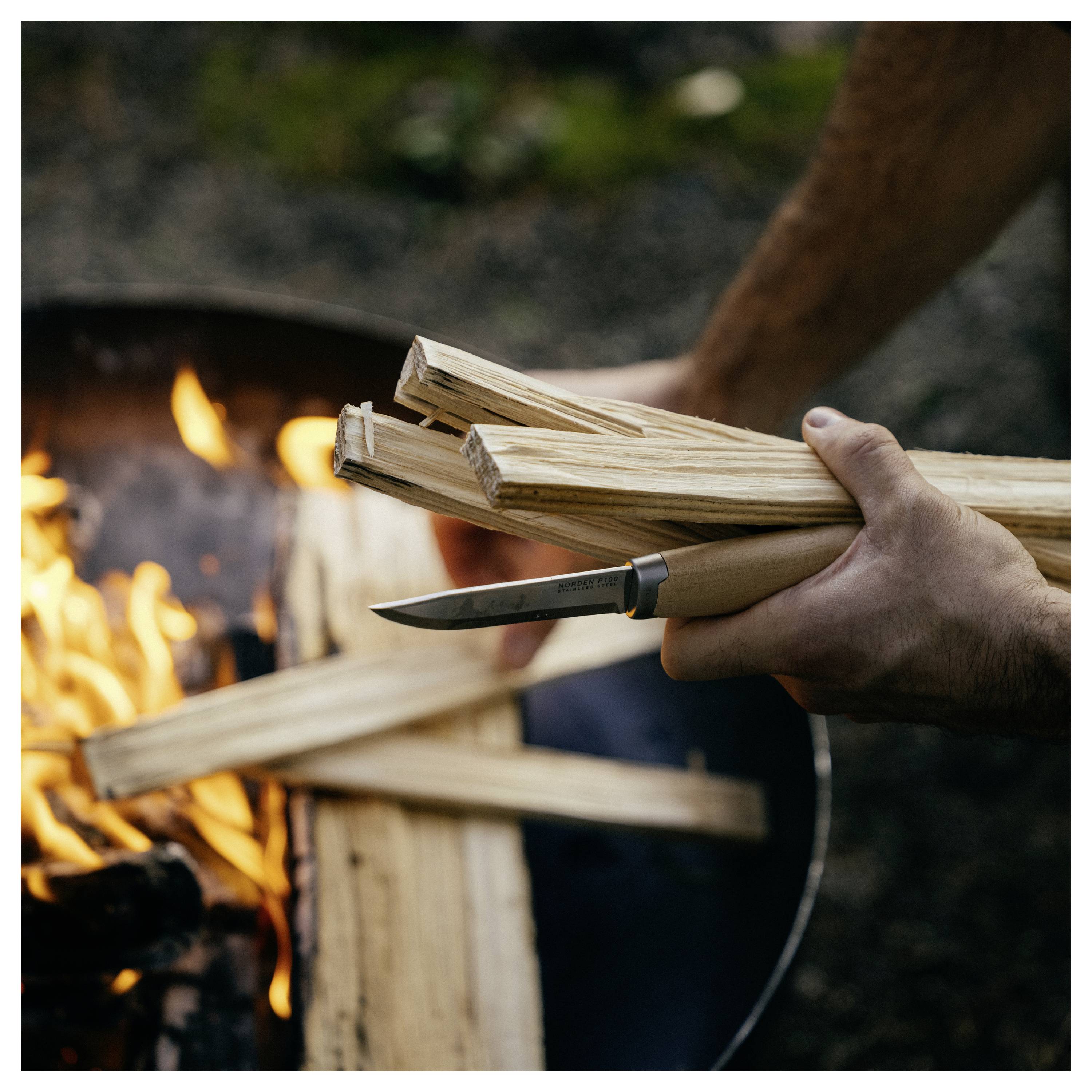 Hände halten gespaltenes Brennholz und ein Messer, während Anmachholz in ein brennendes Lagerfeuer im Freien gelegt wird.