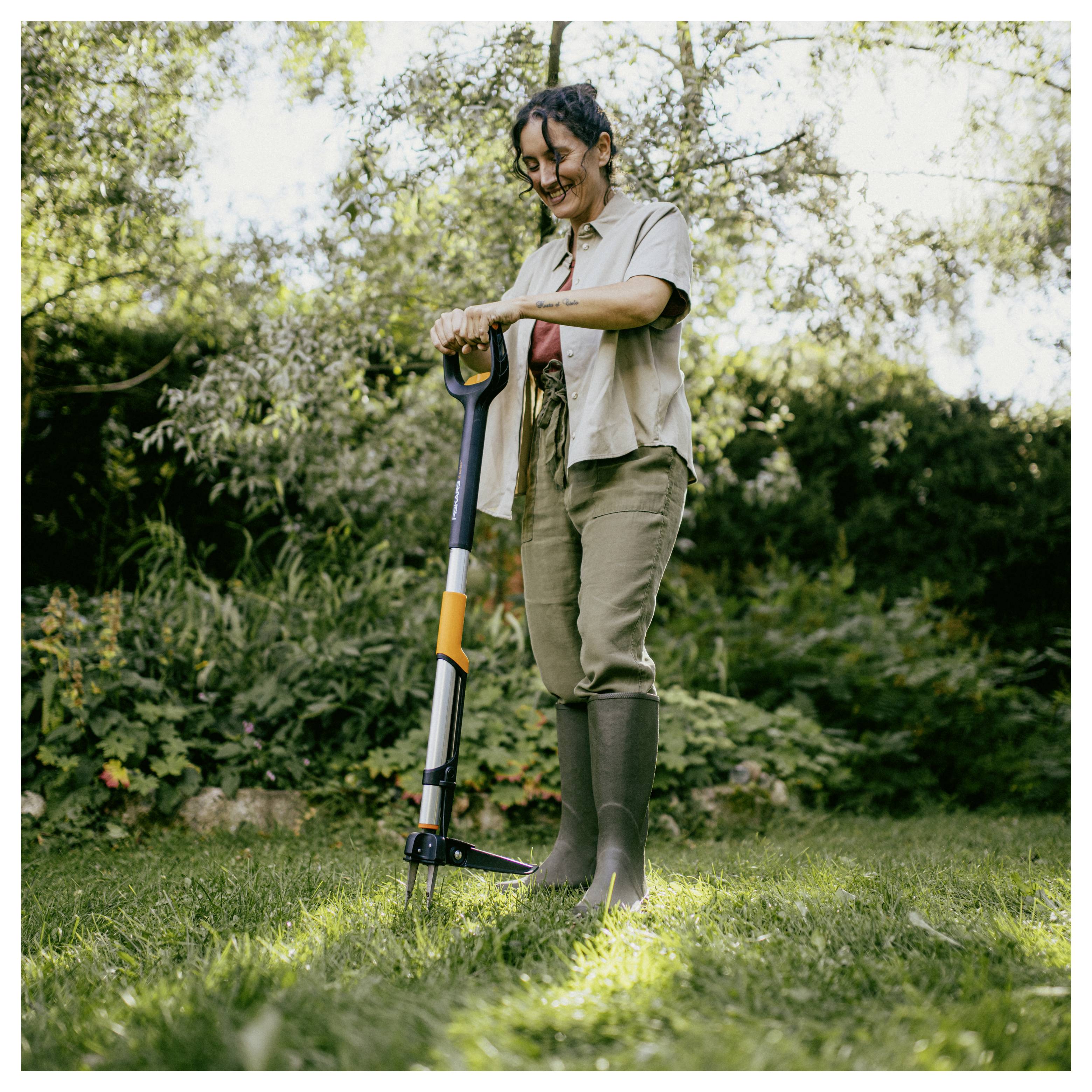 Frau, die an einem sonnigen Tag im Garten mit einer Schaufel arbeitet, steht auf Gras mit Bäumen und Sträuchern im Hintergrund.