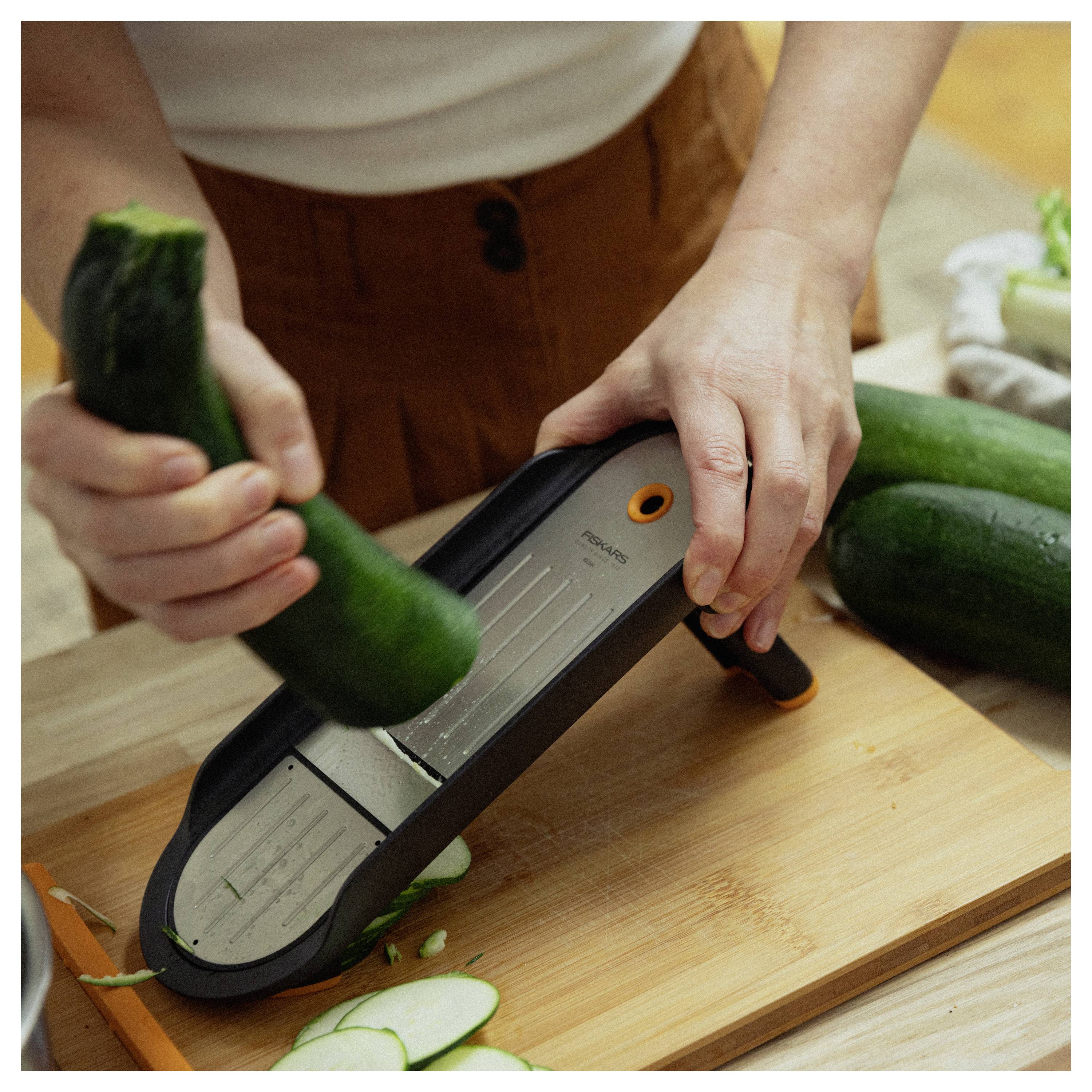 Eine Person schneidet eine Gurke auf einem Mandolinenreißer über einem Holzschneidebrett, mit weiteren Gurken in der Nähe.