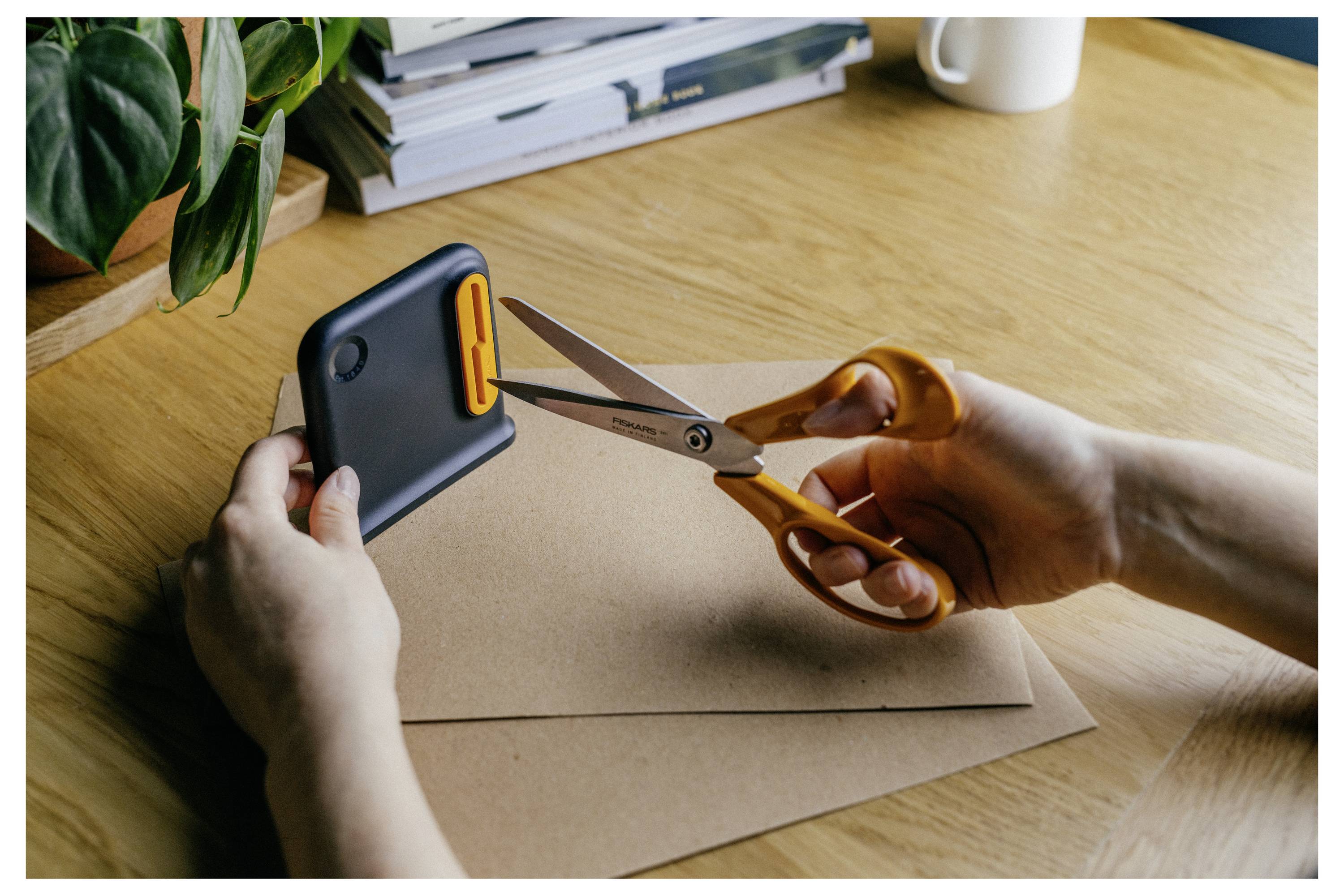 Hände schneiden mit einer Schere ein Stück Pappe ab, während sie ein Smartphone halten, auf einem Schreibtisch mit einer Pflanze und Papieren im Hintergrund.