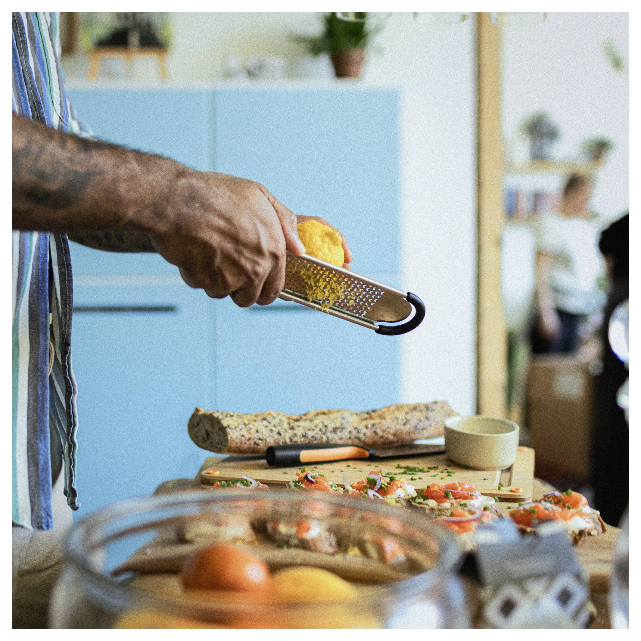 Eine Person reibt Orangenschale über ein Schneidebrett mit appetitlichen Häppchen und Zutaten, die in einer Küche zum Kochen vorbereitet werden.