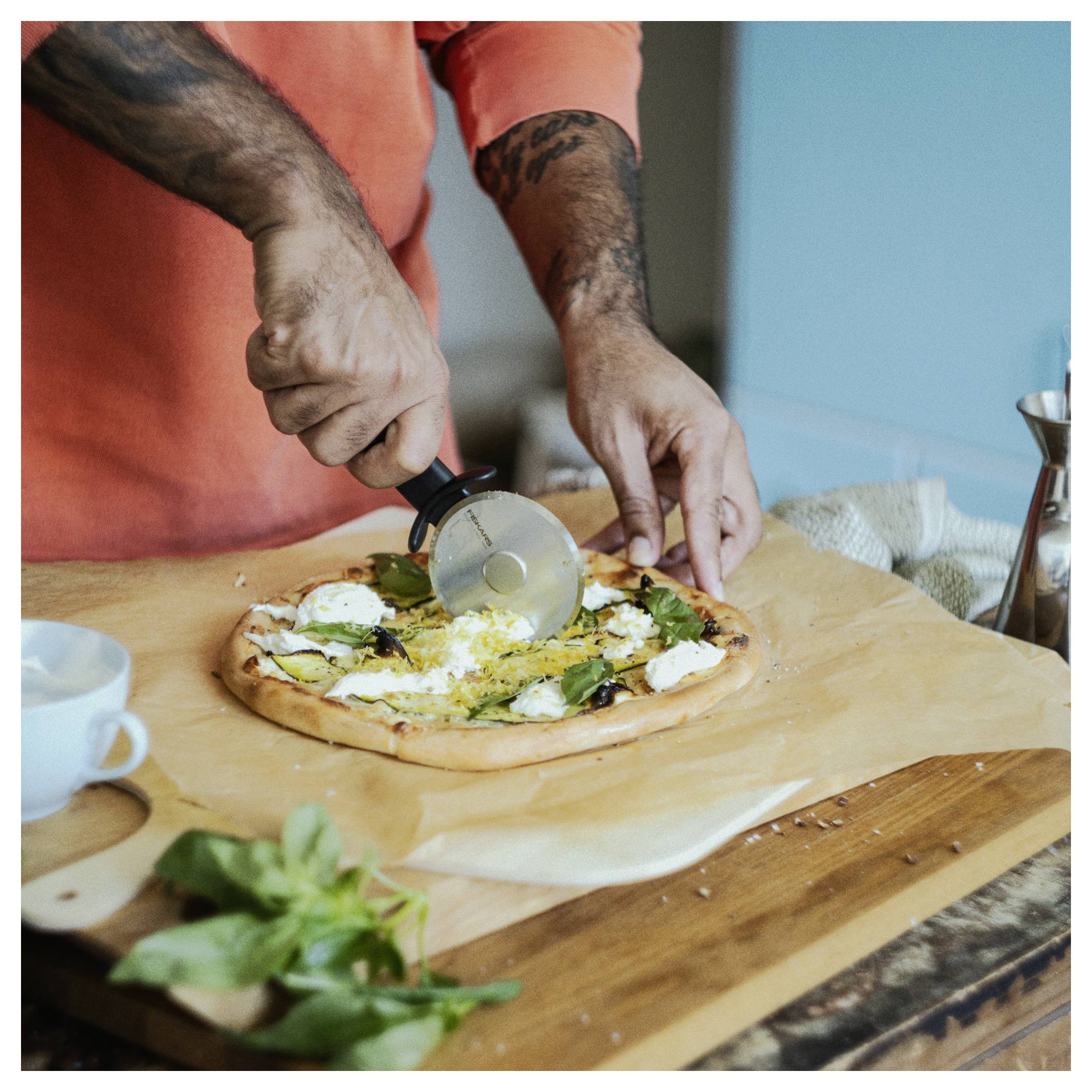 Eine Person in einem orangefarbenen Hemd schneidet eine frische Pizza mit einem Pizzaschneider auf einem Holzbrett, mit Basilikum und einer Tasse in der Nähe.