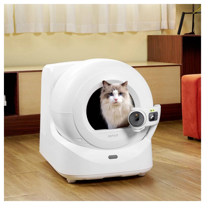 Eine langhaarige Katze sitzt in einer weißen selbstreinigenden Katzentoilette mit angebrachter Kamera auf einem Holzboden in einem Wohnzimmer.