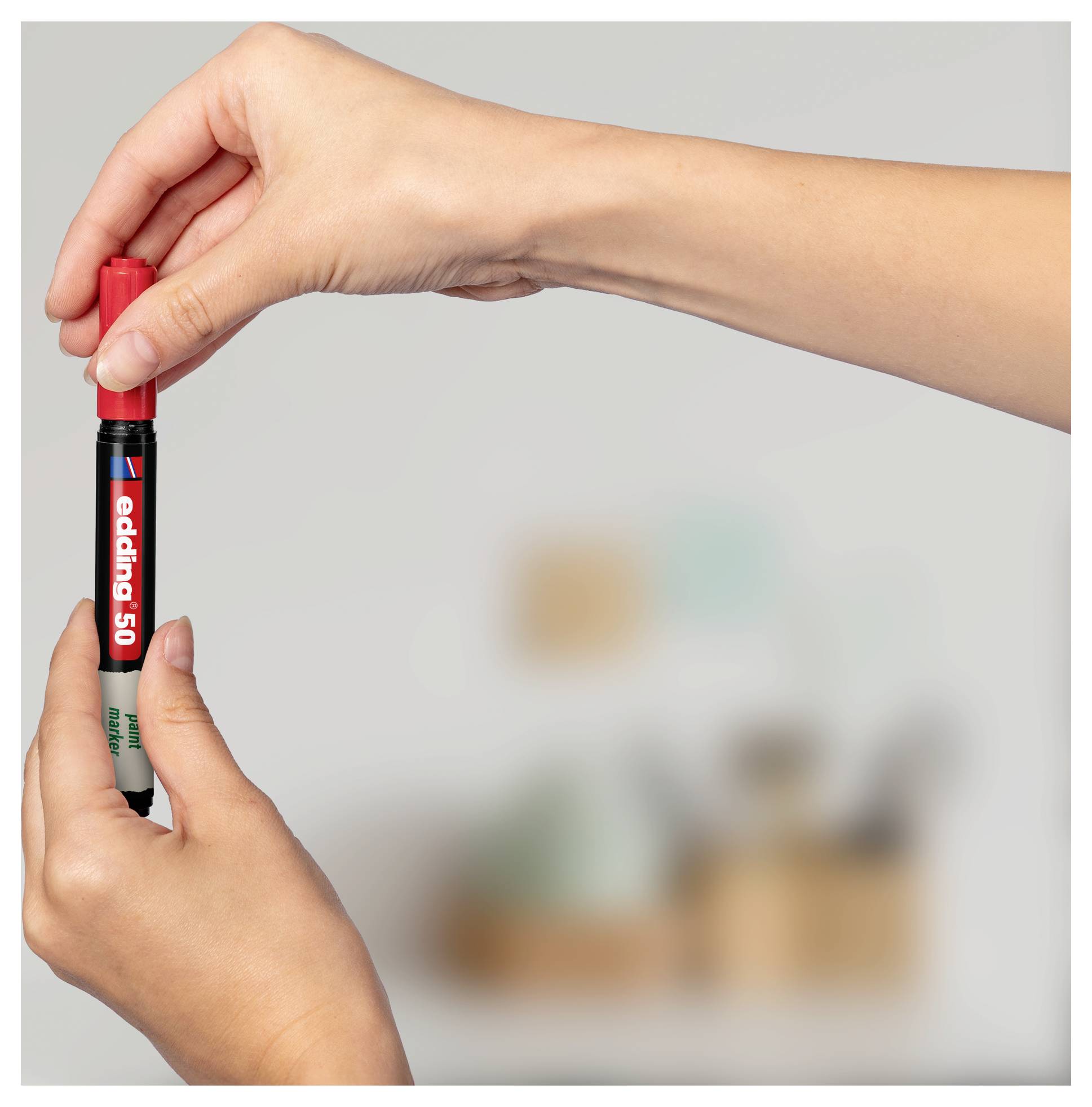 Hände, die einen roten Edding 50 Marker-Stift aufrecht gegen einen unscharfen Innenraum-Hintergrund halten.