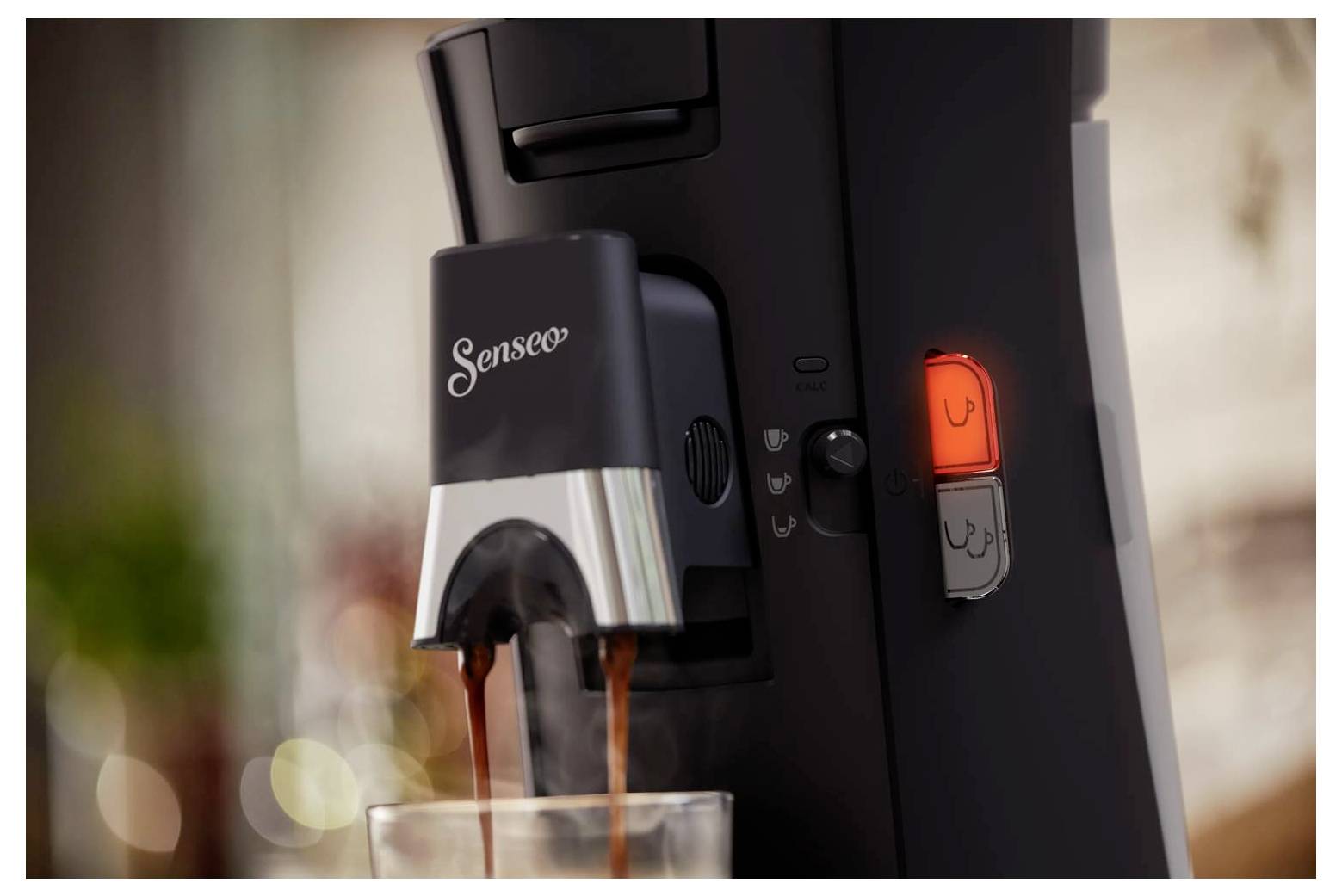 Nahaufnahme einer Senseo-Kaffeemaschine, die Kaffee in ein Glas abgibt, mit dem beleuchteten Brühknopf, der an der Seite sichtbar ist.