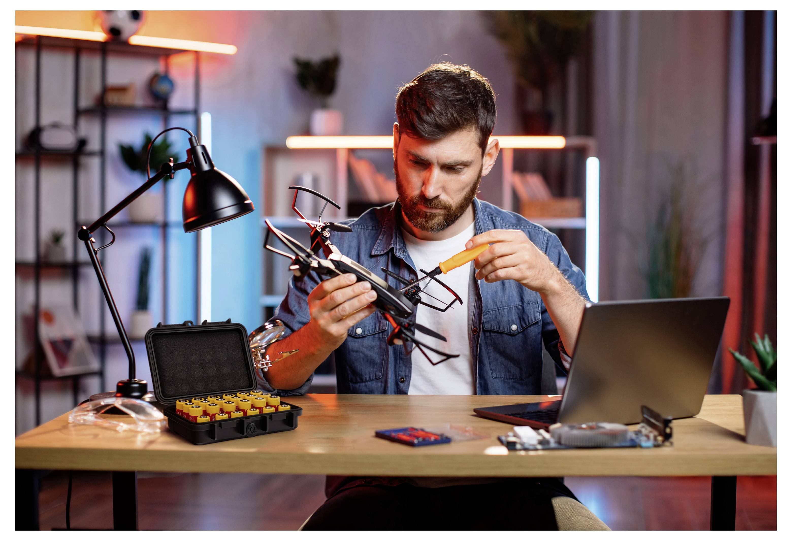 Ein Mann untersucht und repariert eine kleine Drohne an einem Schreibtisch, mit Werkzeugen, einem Laptop und einem offenen Ersatzteilkasten in einem werkstattähnlichen Raum.