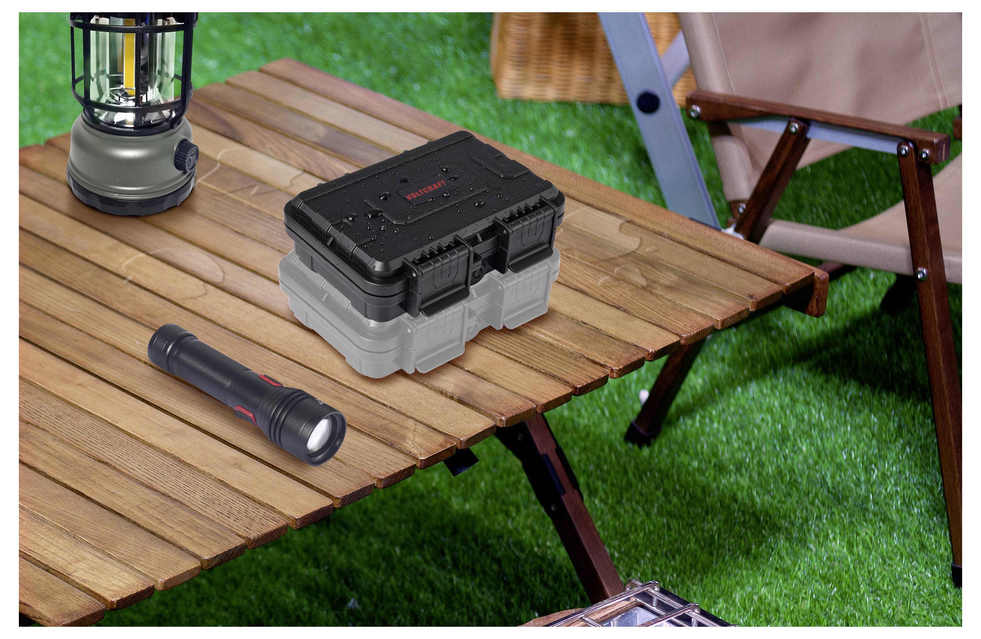 Campingtisch mit einer Laterne, Taschenlampe und robustem Aufbewahrungskoffer auf einem Holz-Outdoor-Tisch neben einem Klappstuhl auf Gras.