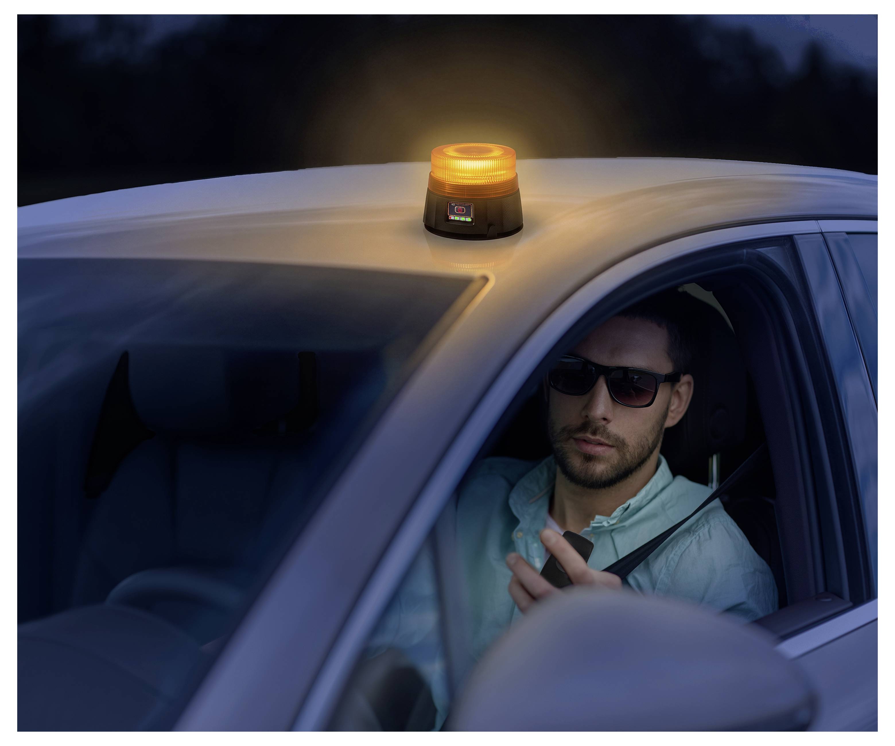 Mann in einem Auto bei Dämmerung mit einem blinkenden bernsteinfarbenen Warnfeuer auf dem Dach, der ein Funkgerät oder Telefon in der Hand hält.
