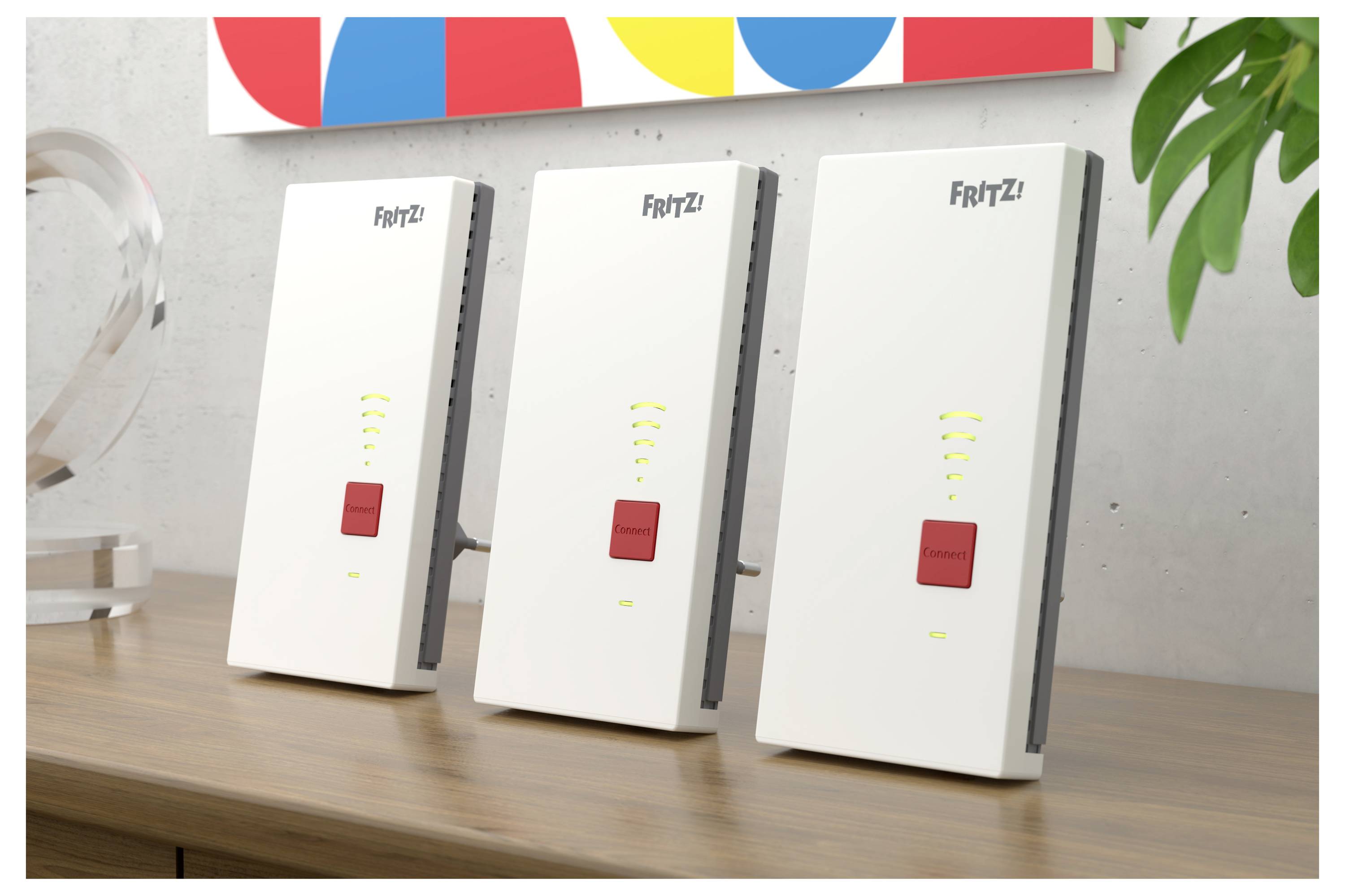 Drei weiße FRITZ! Wireless-Geräte stehen aufrecht auf einem Holzschreibtisch, jedes mit einer roten Taste und Signalleuchten, neben einer Pflanze.